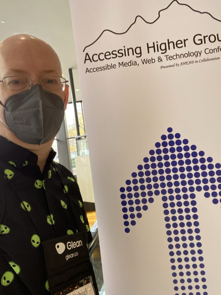 Me standing beside an AHG poster in the hotel lobby. I'm wearing a black shirt with green alien heads and a grey KN95 mask.