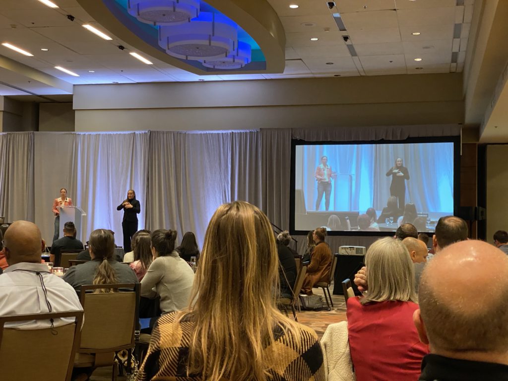 Elsa Sjunneson on stage at the conference keynote. An ASL interpreter stands beside her. Both are also shown on a large video screen to one side of the stage.