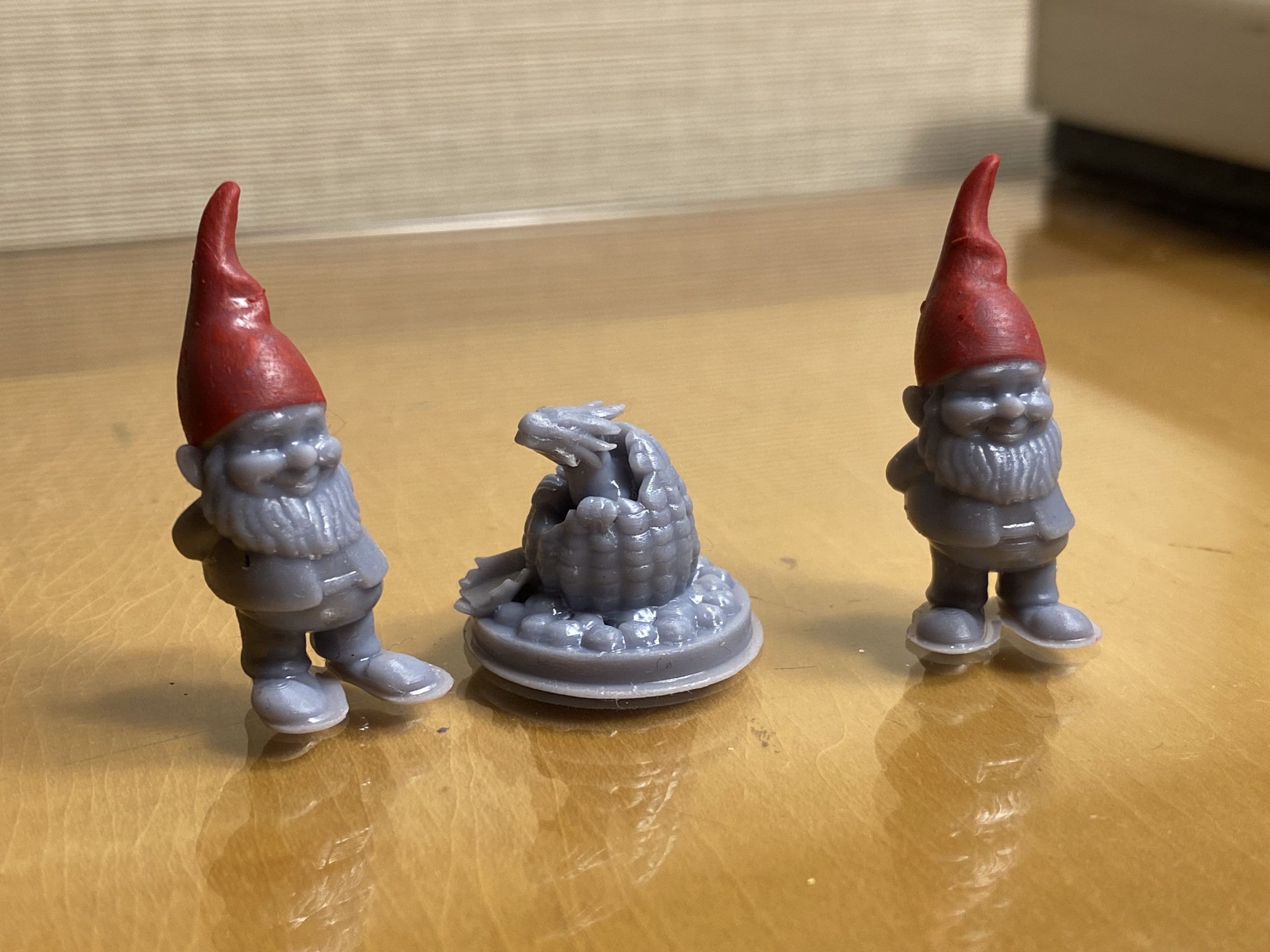 Two small gnomes and a dragon hatching from an egg, all 3D printed in grey, but the gnomes have had their hats painted red.
