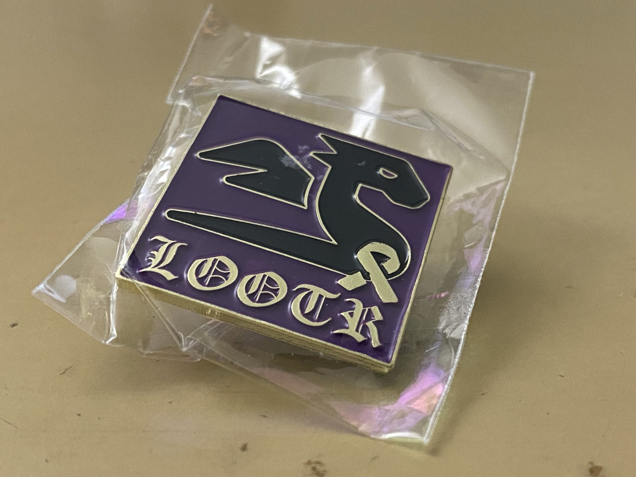 A small square purple enamel pin with a stylized black dragon and the letters “LOOTR” in fancy type.