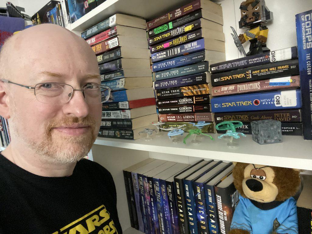 Me in front of a bookshelf full of Star Trek books and a few related toys.