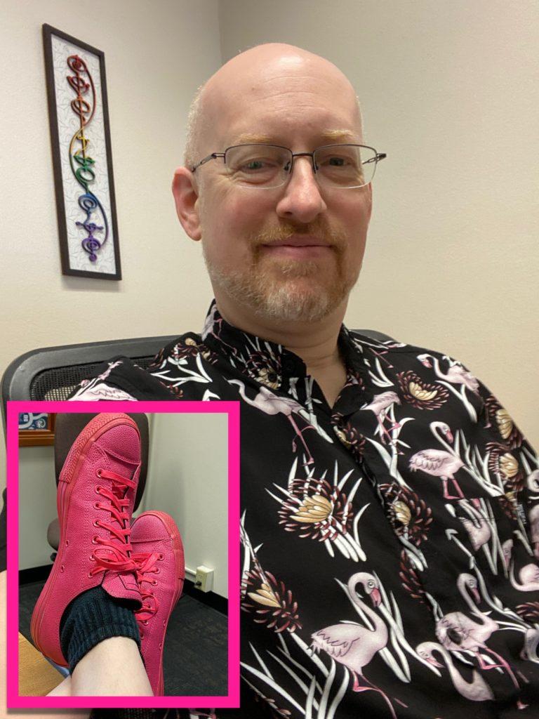 Me sitting in my work office, wearing a black shirt-sleeve button-up shirt with a pattern of pink flamingoes and tropical flowers. Behind me on the wall is a rainbow IDIC Vulcan calligraphy plaque. An inset photo shows my bright pink Converse. 