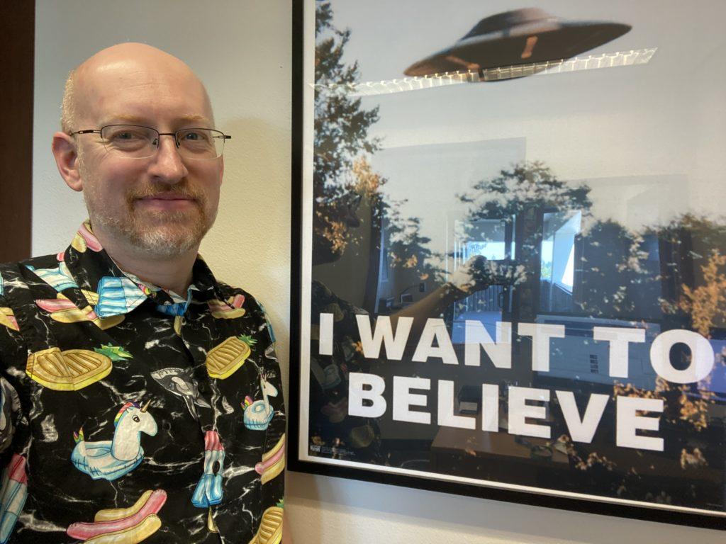 Me in my office at work, standing next to a reproduction of Mulder’s ‘I want to believe’ poster from The X-Files, wearing a black short-sleeve button-up with a print of unicorn, pineapple, popsicle, and hot dog inflatable pool floaties.