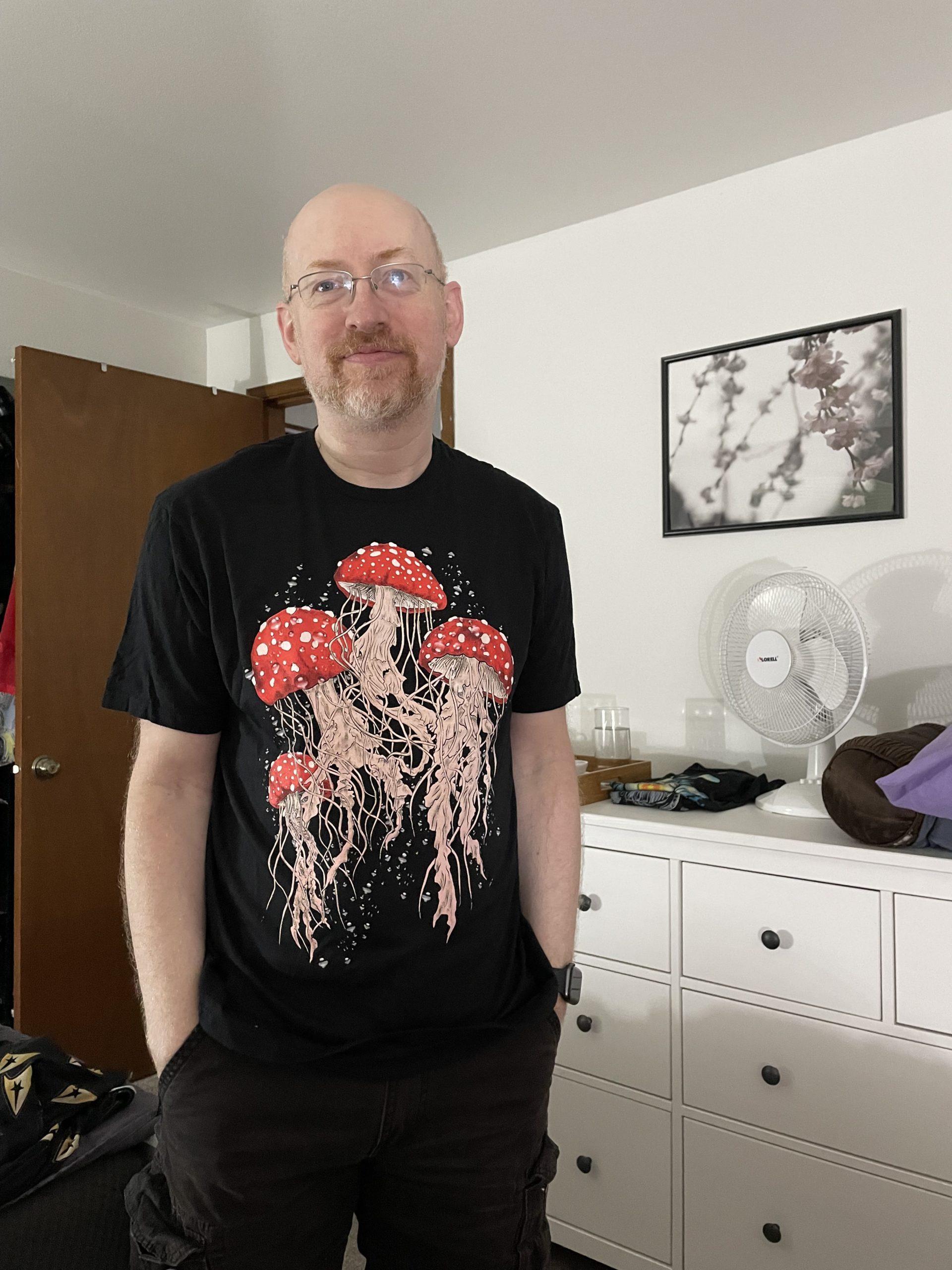 Me standing in our bedroom in front of a white dresser. I'm wearing black shorts and a black t-shirt with a print of jellyfish, but the tops look like amanita muscaria mushrooms, with white-dotted red caps.