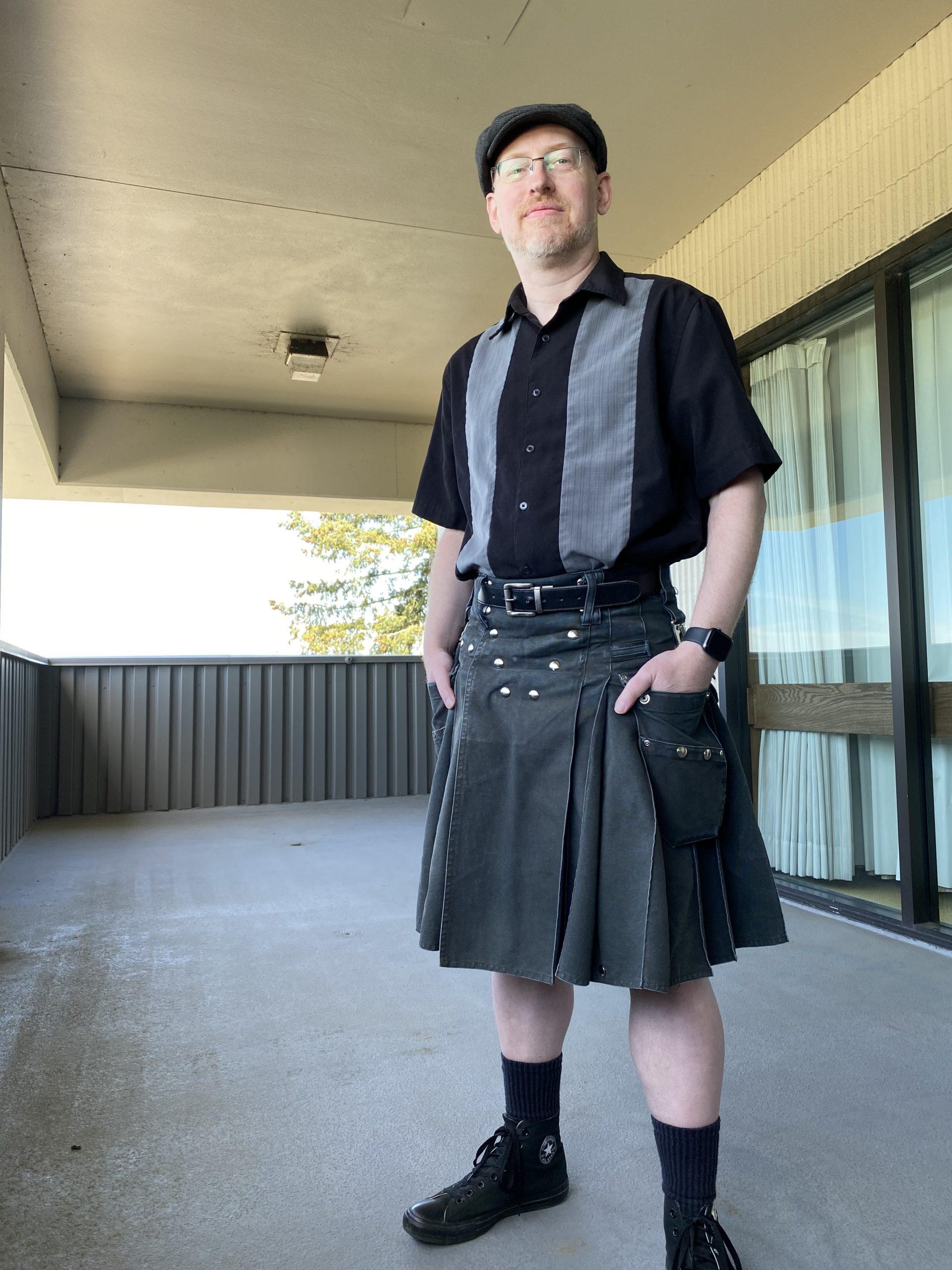 Me standing on an outside balcony wearing a grey cap, black short-sleeve button-up shirt with thick grey stripes, a black utility kilt, black socks, and black Converse.