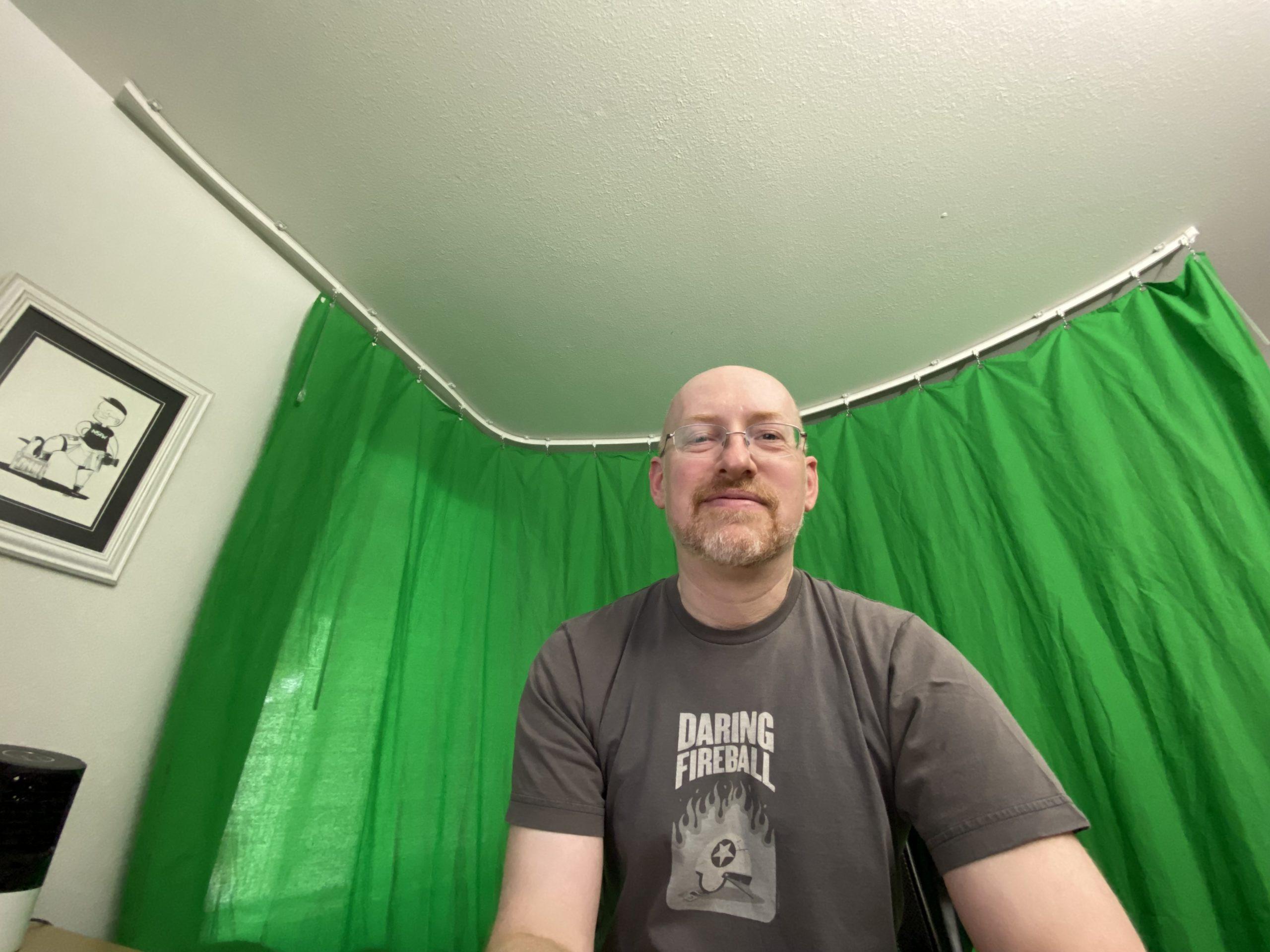 Me wearing a grey Daring Fireball t-shirt in front of a green curtain suspended from the ceiling of my home office.