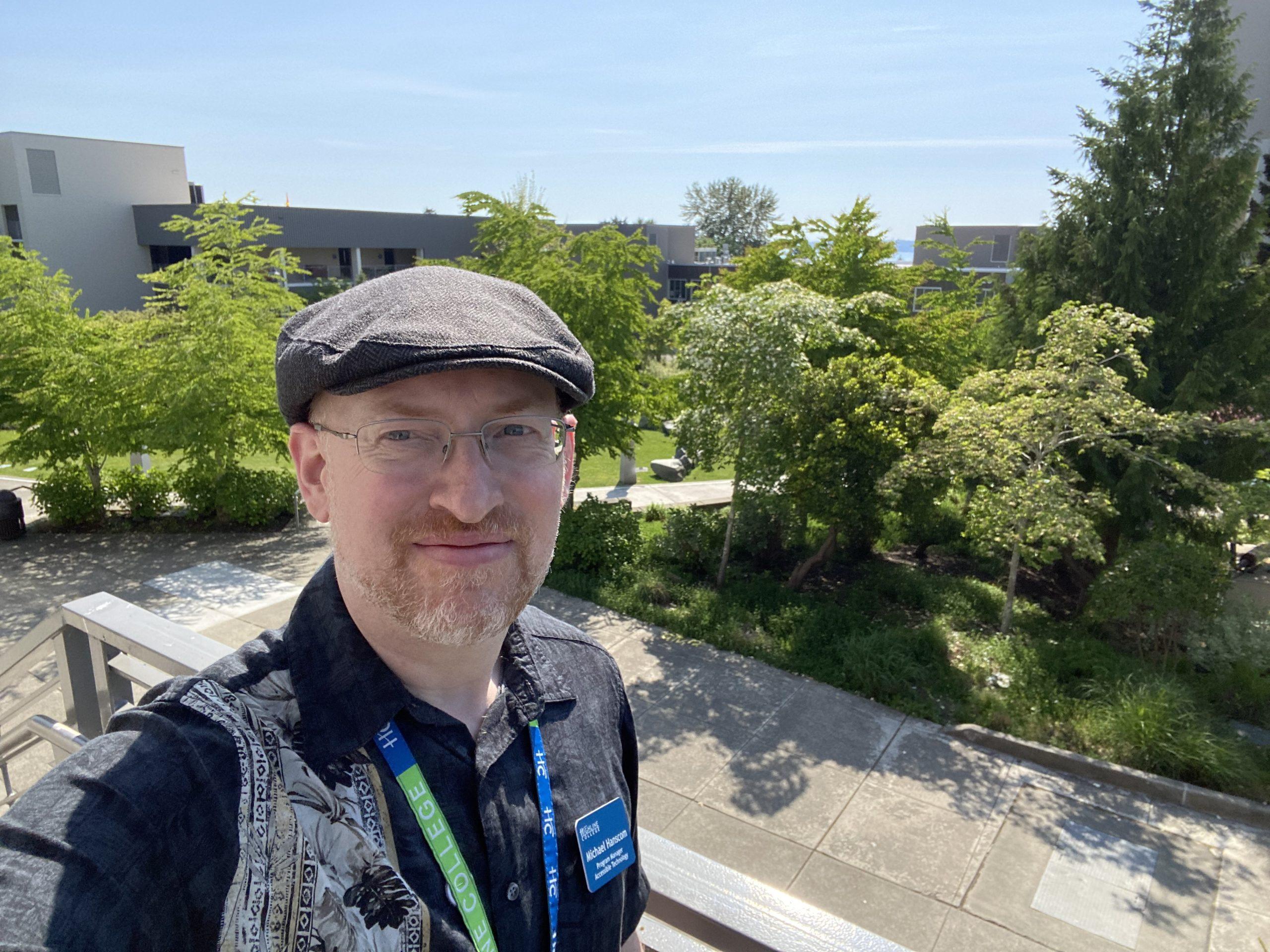 Me standing outside wearing a grey cap and black short-sleeve button-up shirt with a floral pattern on the right half. Behind me is the college campus.