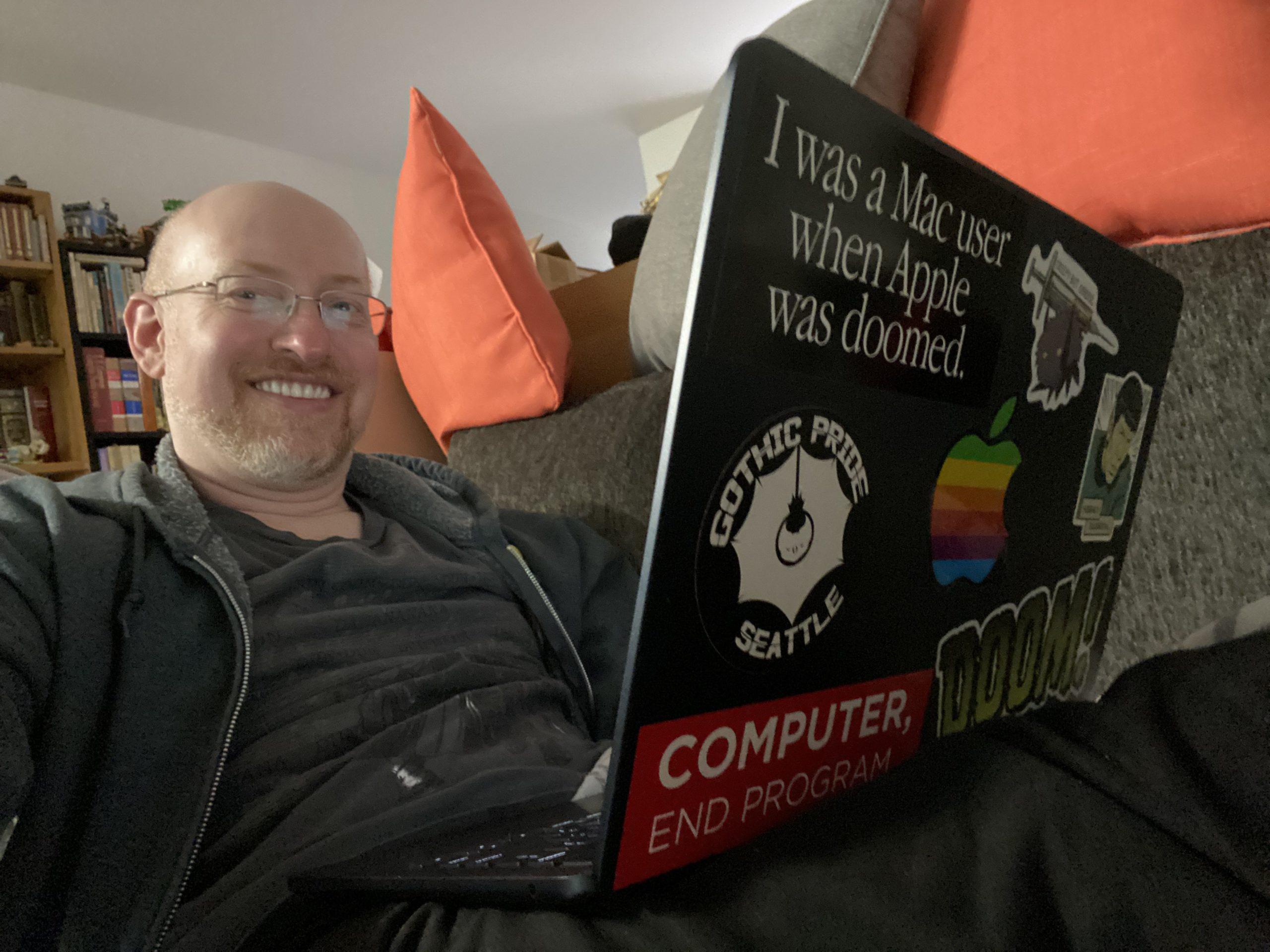 Me kicked back on a dark grey couch with light grey and orange pillows behind me. On my lap is an Apple Macbook Air with a number of stickers on the lid.