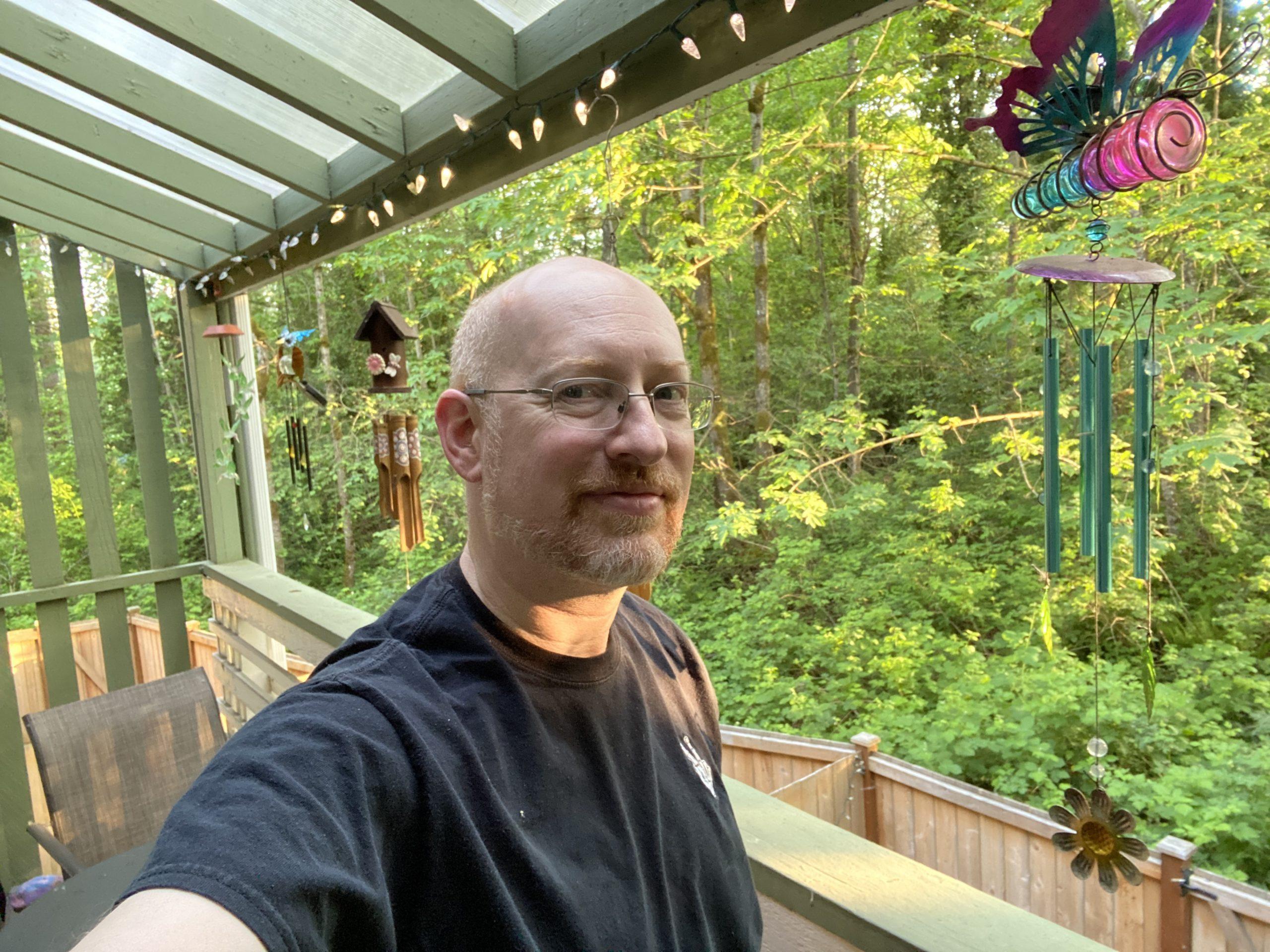 Me standing on a back deck, with several windchimes hanging from the deck roof, and lots of green trees in the background.