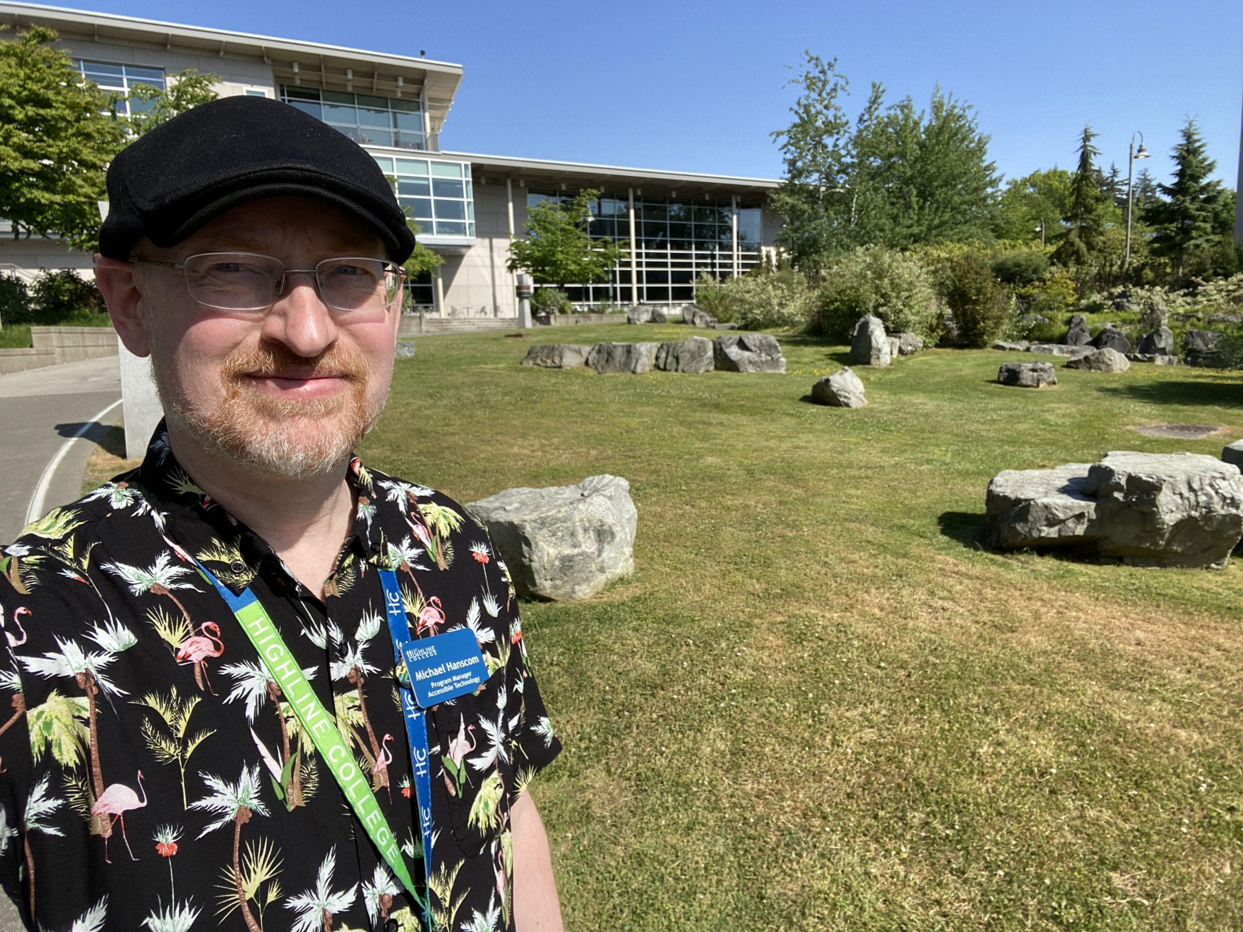 Me, wearing a black short-sleeve button-up shirt with flamingos and tropical plants, standing in front of a green lawn with varied decorative rocks, with trees, blue sky, and a campus building in the background.