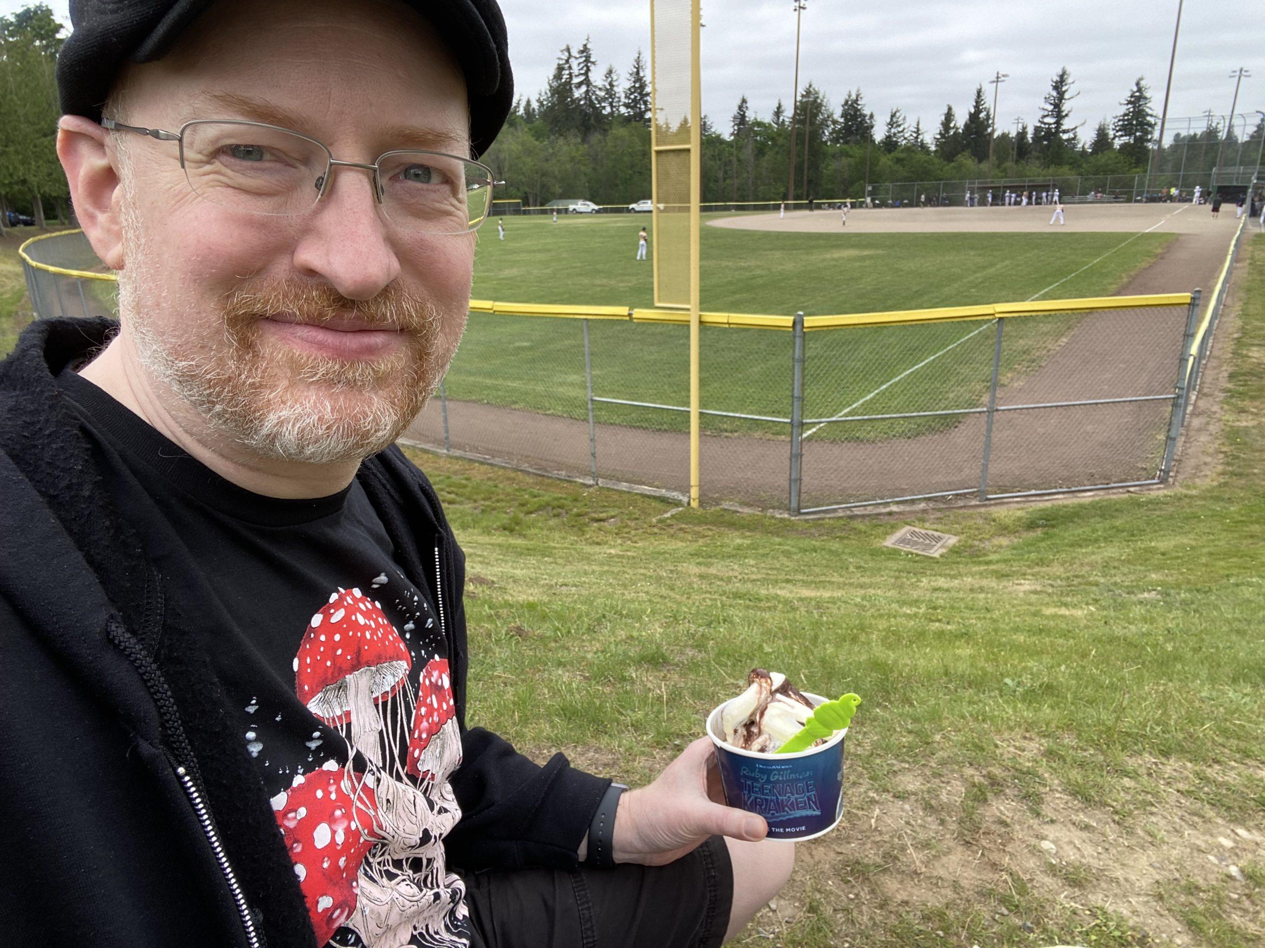 Me sitting outside the fence of a community park baseball diamond, with players visible in the distance. I'm holding a cup of frozen yogurt.