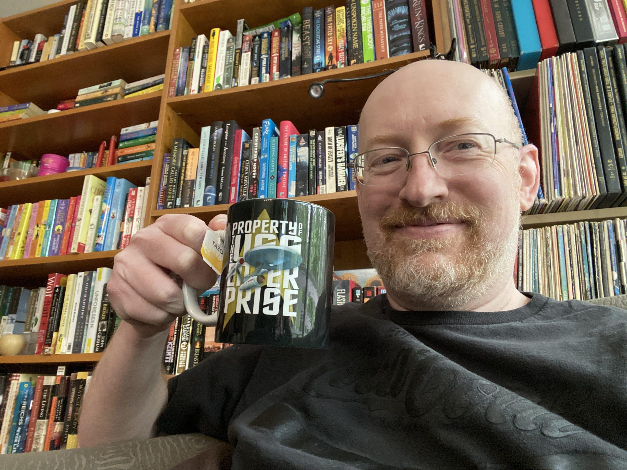 Me in front of several shelves packed full of either books or music LPs. I'm hoding a mug that says "Property of USS Enterprise" with a graphic of the original starship Enterprise.
