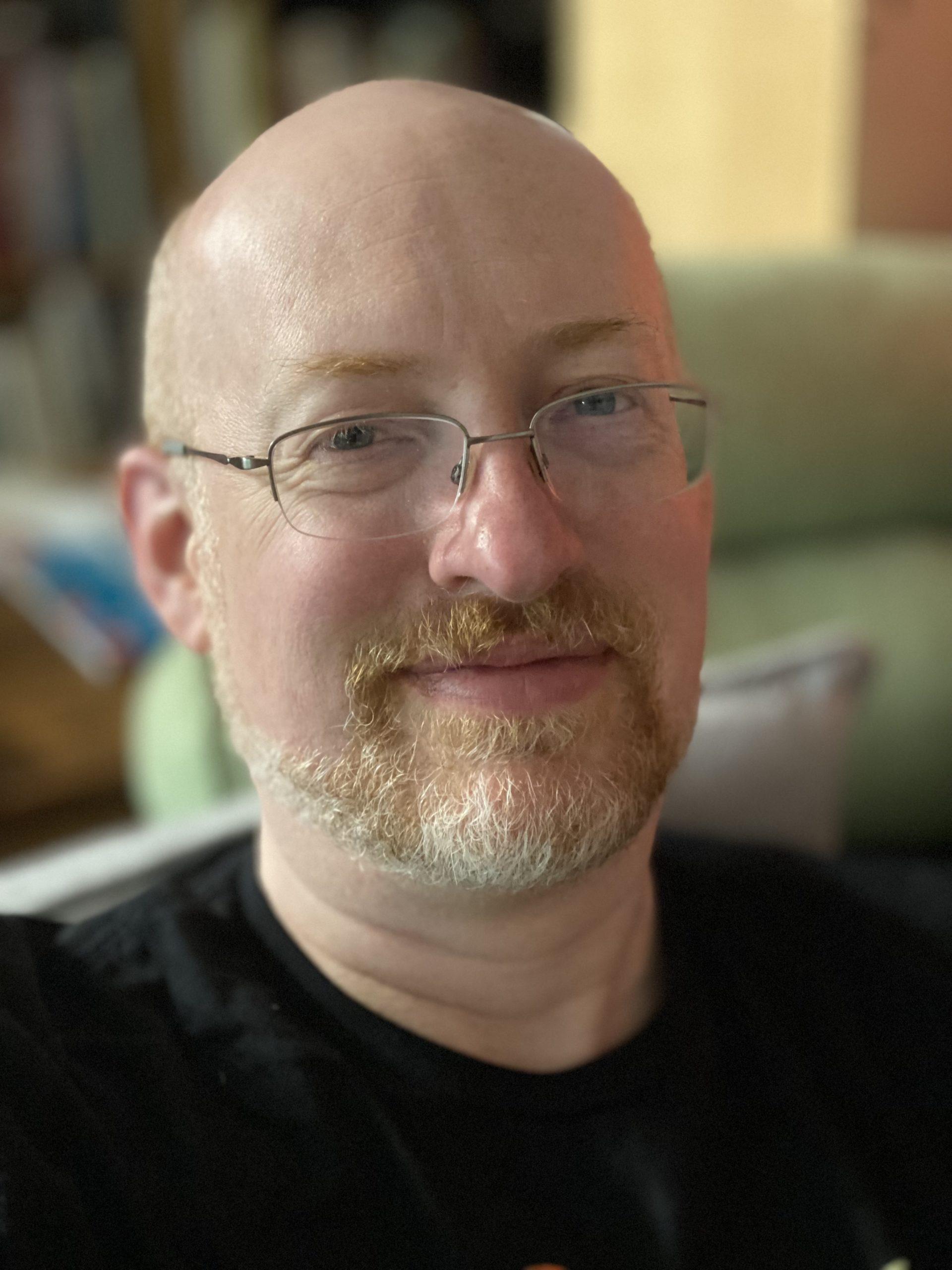 Me, a white, bald man with light red mustache and beard with a lot of white in it around the chin, wearing sillver half-rim wireframe glasses, smiling slightly at the camera.