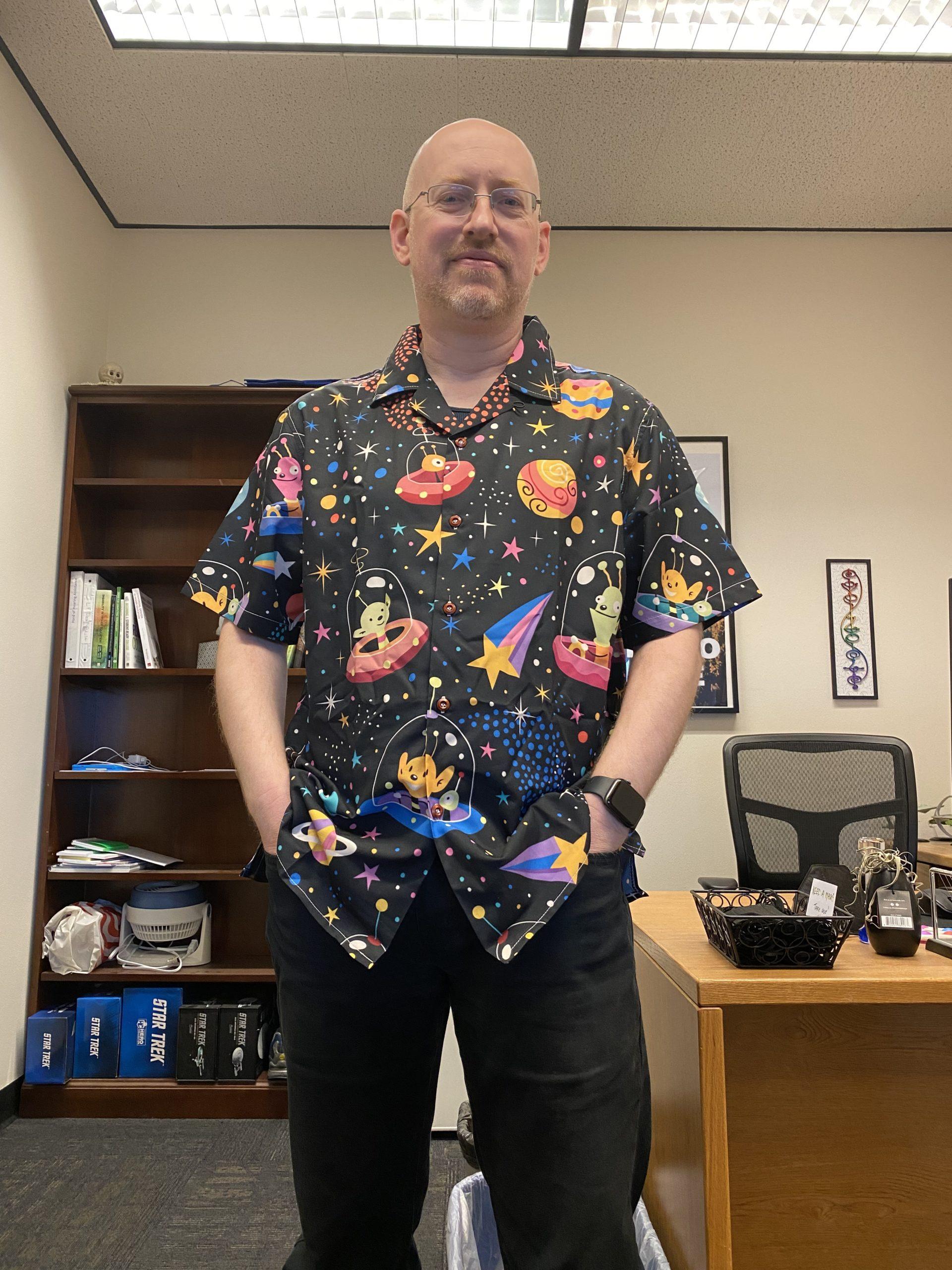 Me standing in my work office, wearing black jeans and a black short-sleeve button-up shirt with muticolored stars, planets, and aliens in UFOs.