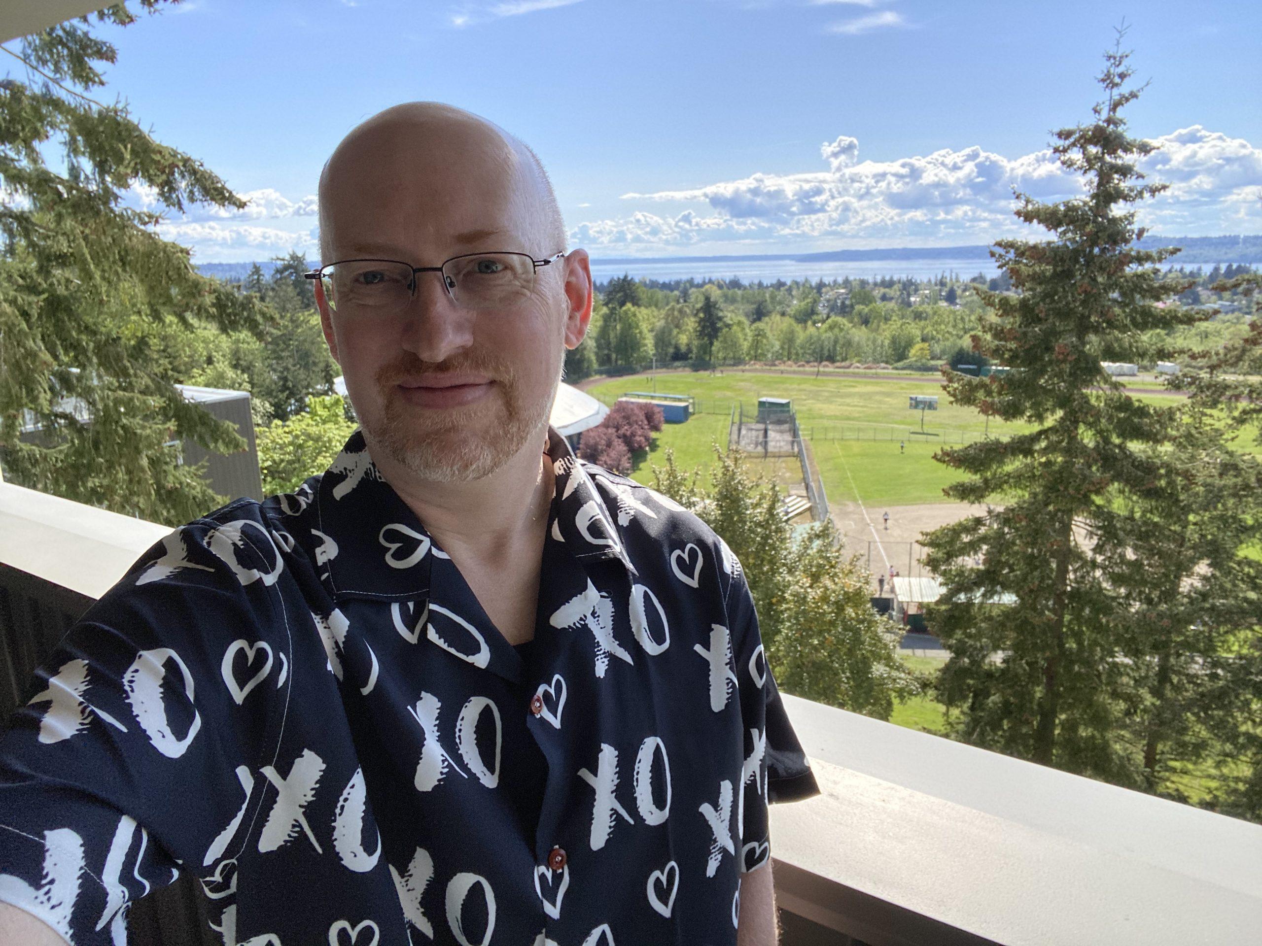 Me standing on a balcony, wearing a black short-sleeve button-up shirt with a design of white Xs and Os. Behind me is blue sky with fluffy clouds, trees and green sports fields, and water in the distance.