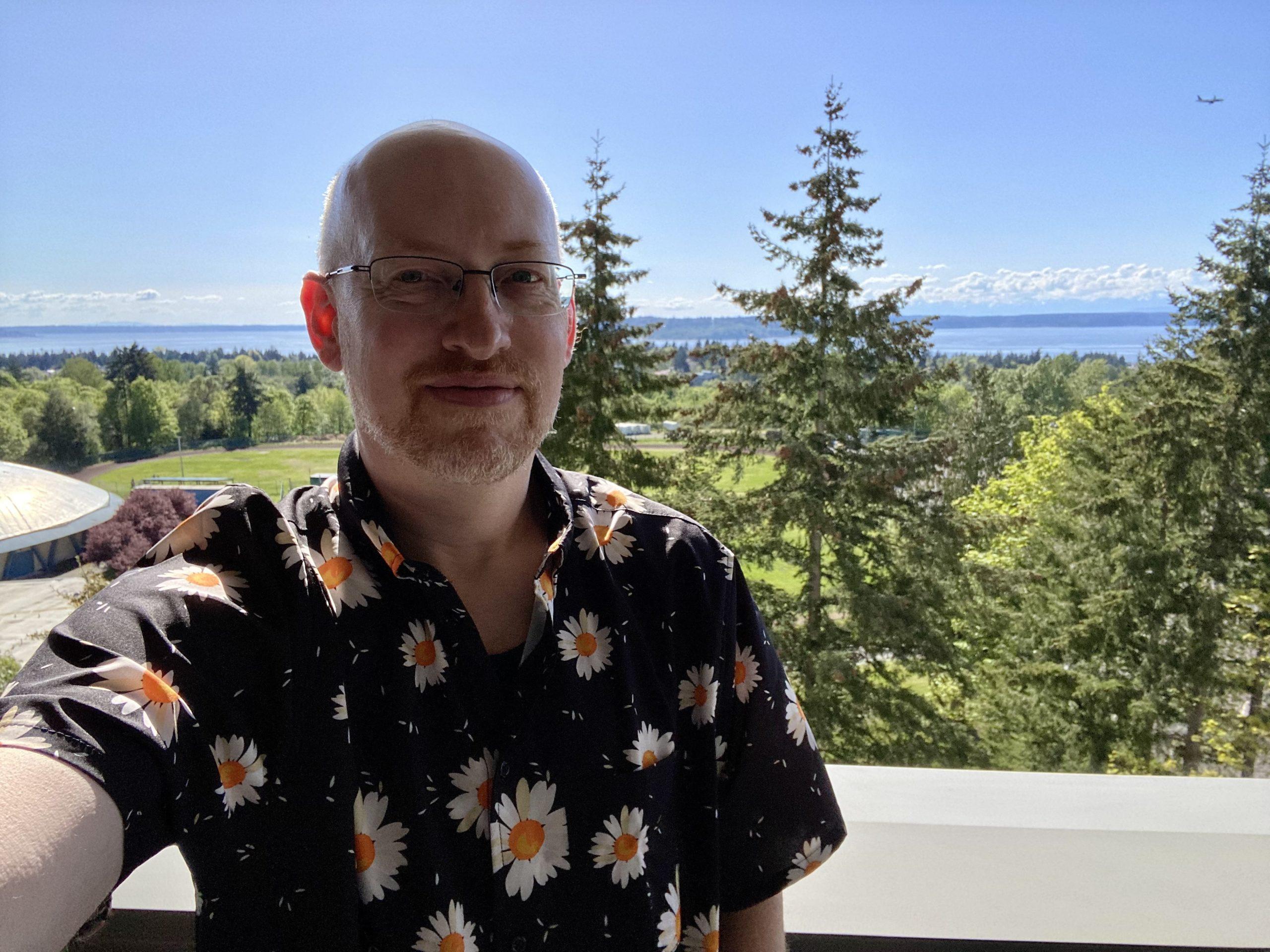 Me wearing a black short-sleeve button-up shirt with daisies printed on it, in front of a view of green trees and grass, water, blue skies, and the Cascade range in the distance.