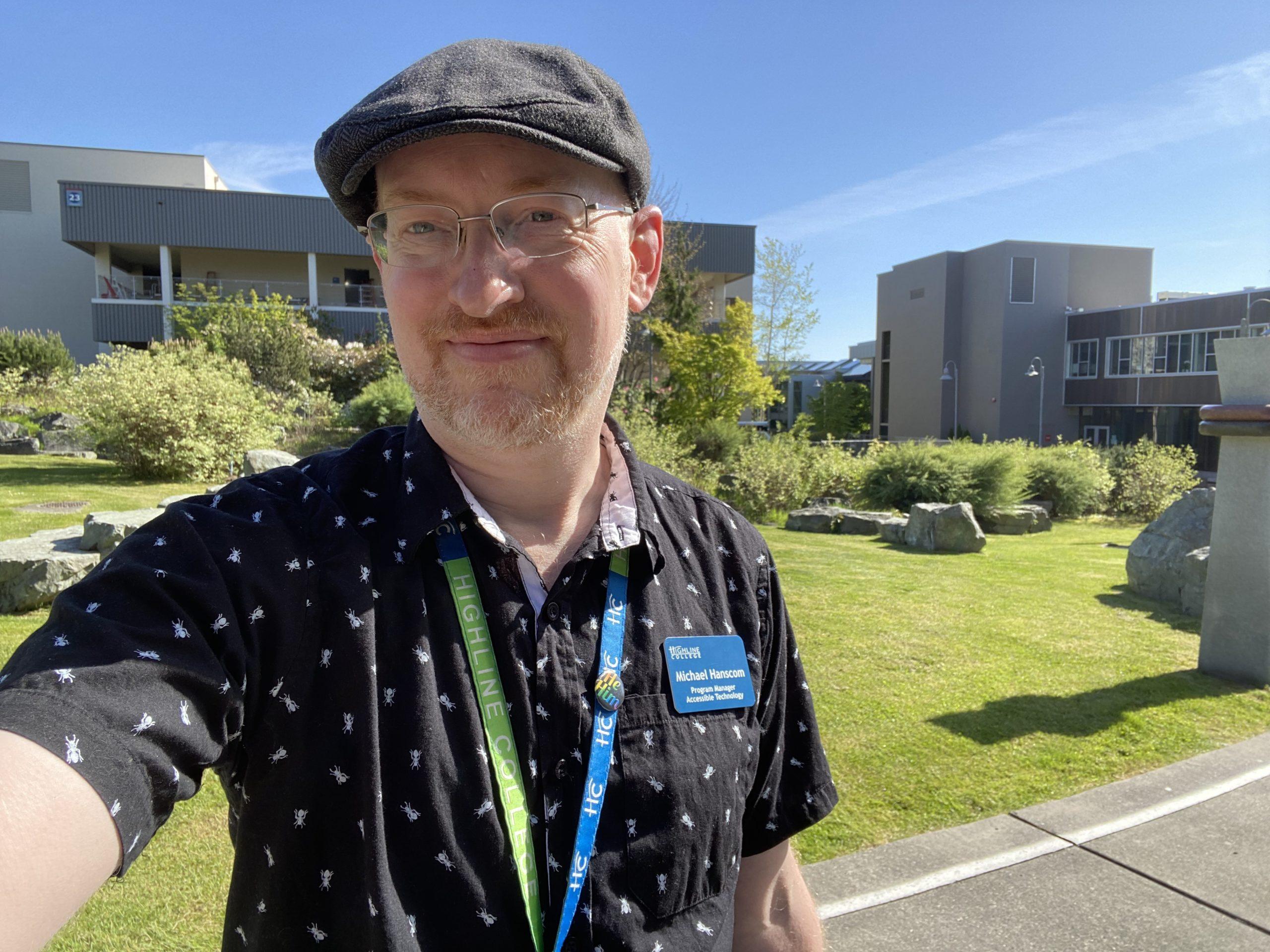 Me on the Highline College campus on a sunny spring day, wearing a grey cap and a black short-sleeve button-up shirt with a pattern of white ants.