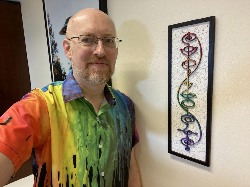 Me wearing a short-sleeve button-up shirt with a print that looks like rainbow paint was poured over and is dripping down the shirt. I'm leaning on my office wall. On the wall next to me is a rainbow-colored laser-cut wood plaque of the Vulcan calligraphy for Kol-ut-shan, or IDIC - Infinite Diversity in Infinite Combinations.