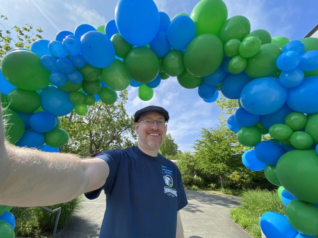 A wide angle selfie of me wearing a Highline College t-shirt standing under an archway of balloons in the Highline blue and green colors.