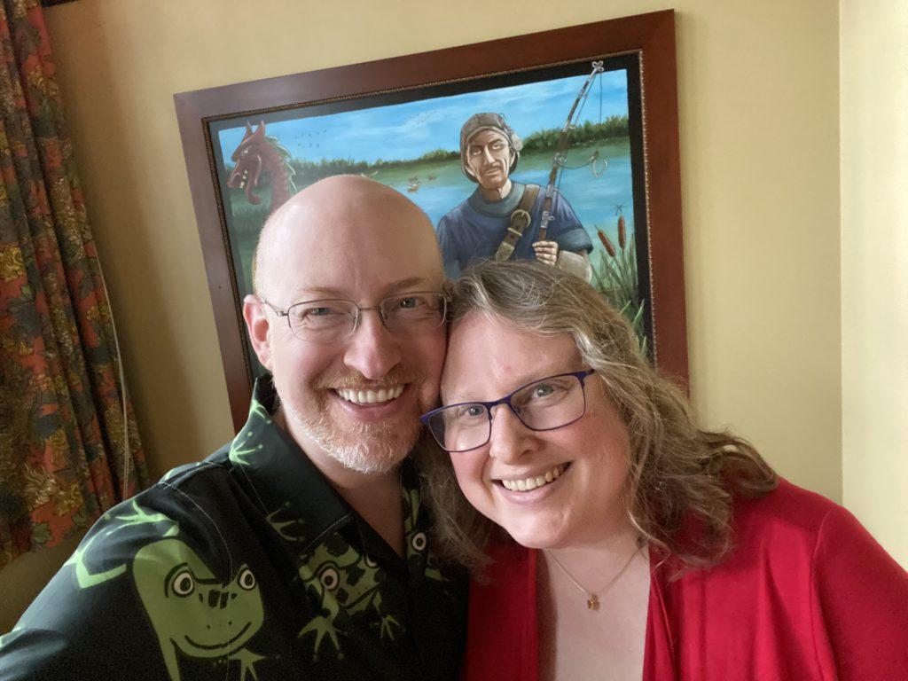 My wife and I, smiling for a selfie in front of a painting of Wilbert Andell Anderson, depicted as a creepy-looking gaunt white man with a brown goatee wearing a blue t-shirt and backwards baseball cap and holding a fishing rod; his eyes are hooded and he has a slight smirk, as if he’s sizing you up and deciding he could easily dispose of you where you wouldn’t be found anytime soon.