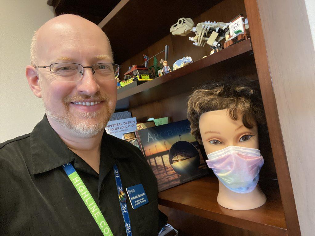 Me standing next to a bookcase; on the shelf beside me is a mannequin head with wavy brown hair wearing a pastel rainbow disposable face mask.