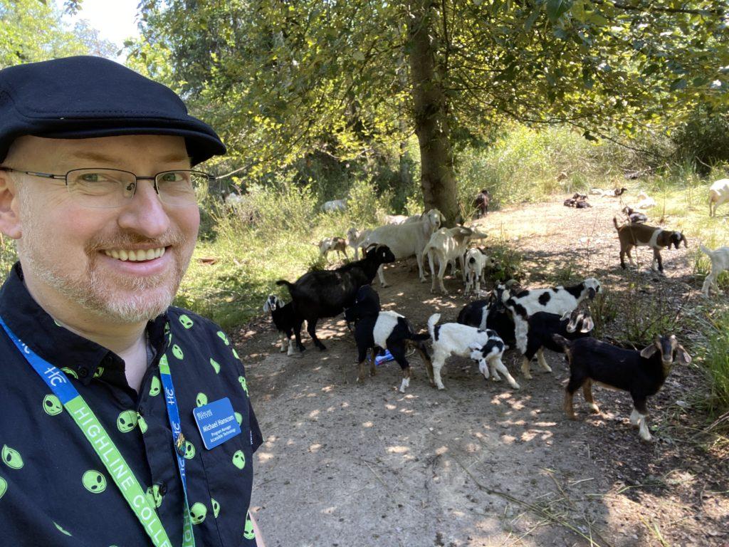 Me wearing a black cap and black shirt with a pattern of green alien heads, standing near a herd of goats munching on undergrowth in a wooded area.