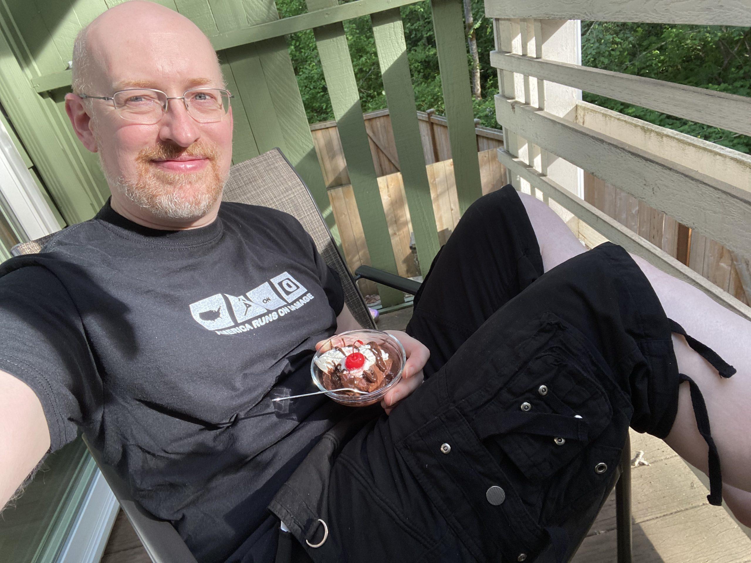 Me sitting on our back deck, wearing black cargo shorts and t-shirt, holding a bowl of chocolate pudding with whipped cream and a maraschino cherry.