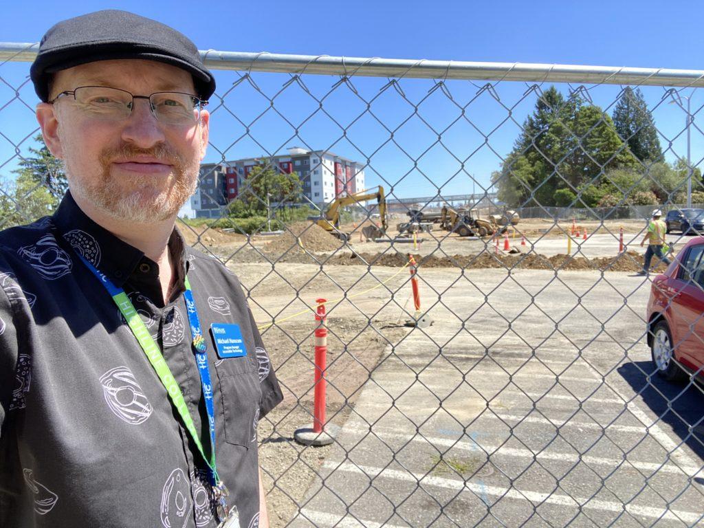 Me standing outside in front of a chain-link fence. Behind the fence is an active construction area, with workers and heavy equipment visible. In the far distance are the tracks for an in-construction light rail station.