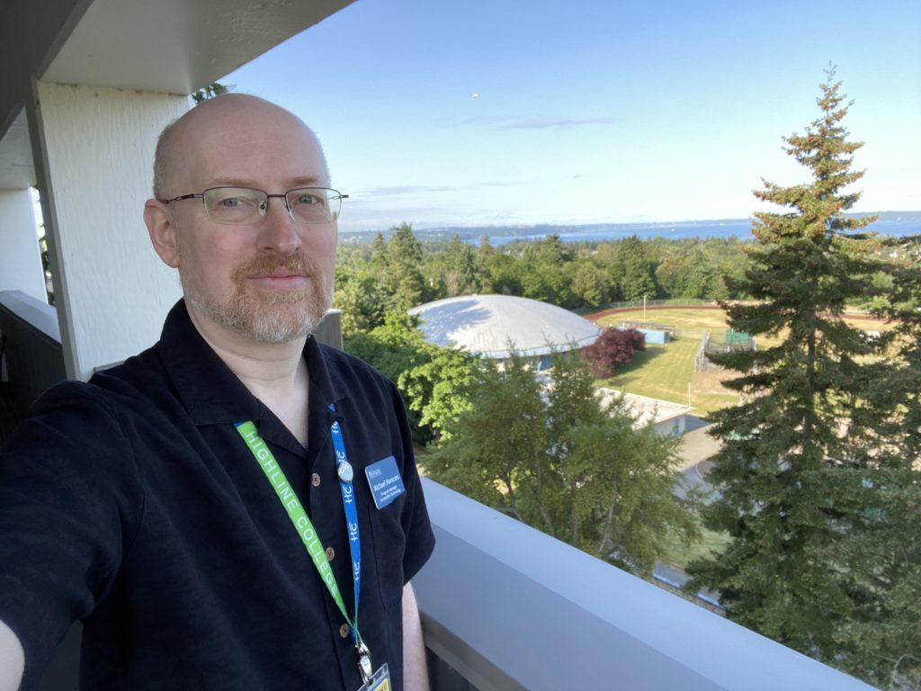 Me on my office balcony, with trees, a sports field, water, and blue skies in the background, wearing a plain black short-sleeve button-up shirt.