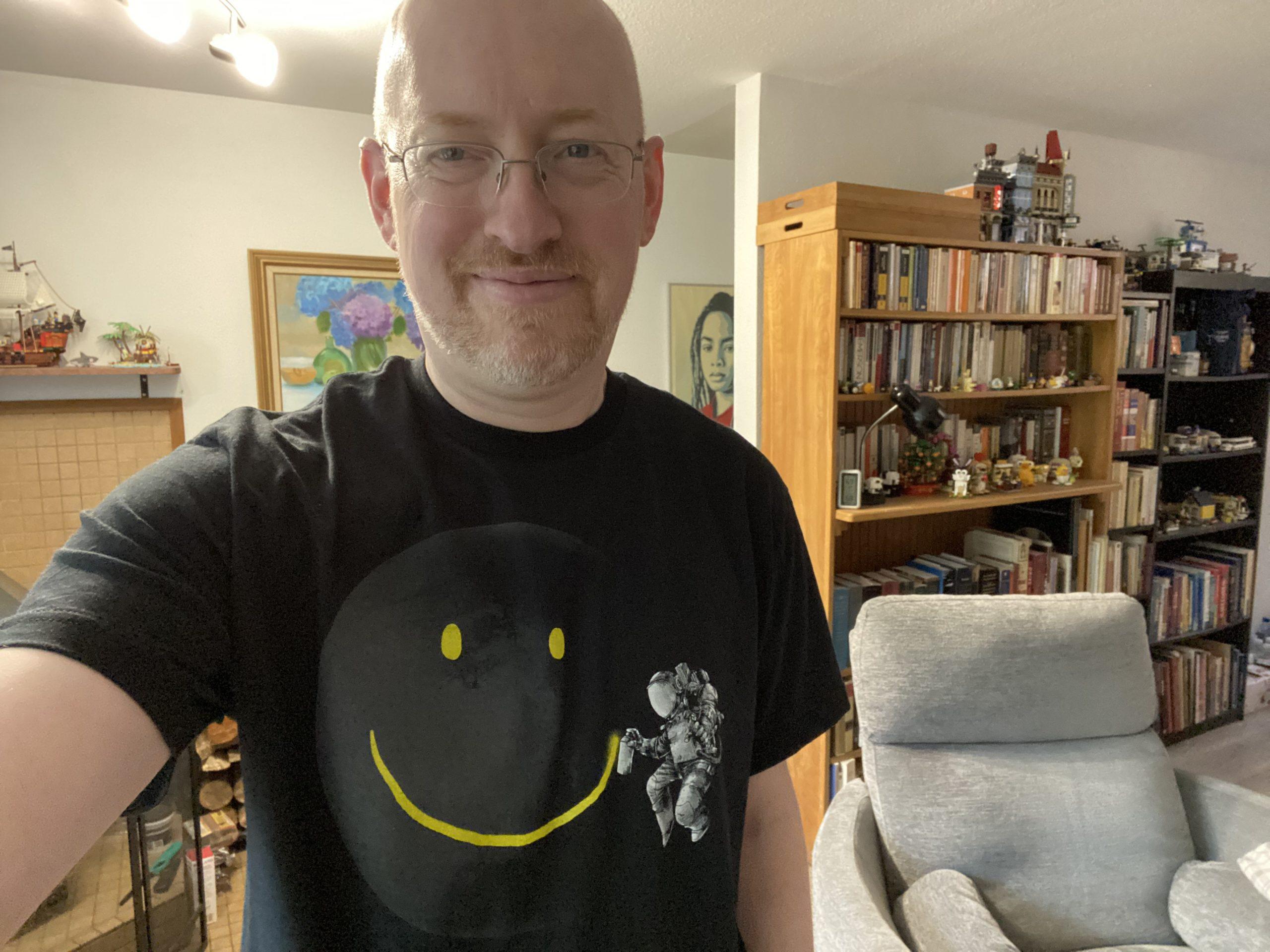 Me standing in my living room, with shelves of books visible behind me. I'm wearing a black t-shirt with a graphic of an astronaut spray-painting a happy face smile on the moon.