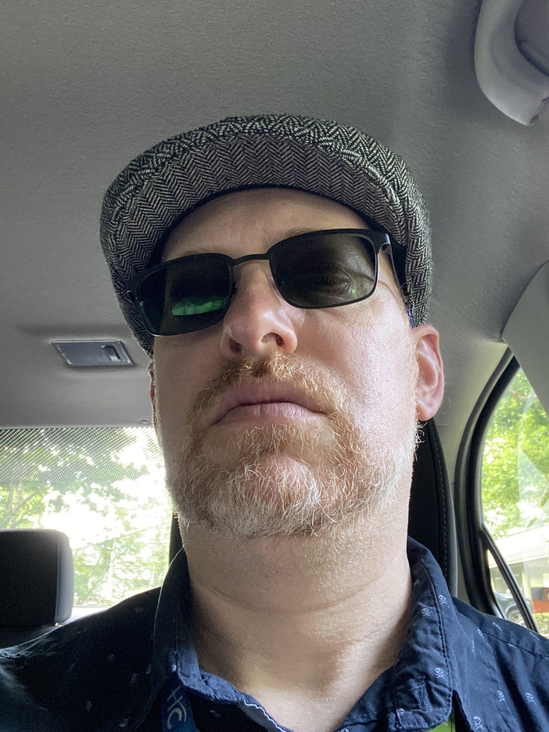 A selfie of me, a 50-year-old white man, sitting in the driver's seat of my car, unsmiling, wearing a hat and sunglasses, not smiling, shot from slightly below.