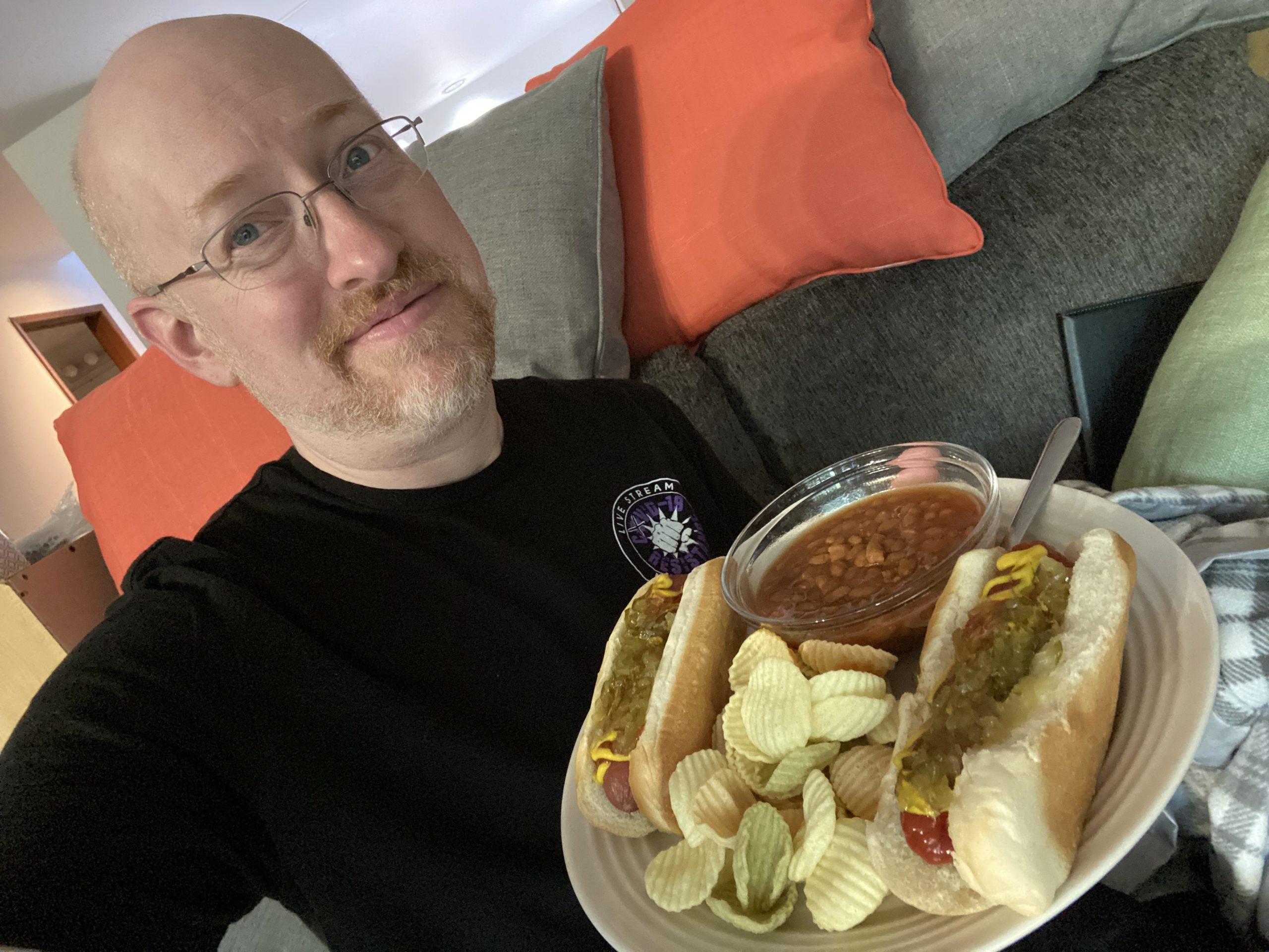 Me in my living room with an unimpressed expression, holding a plate of hot dogs, beans, and chips.