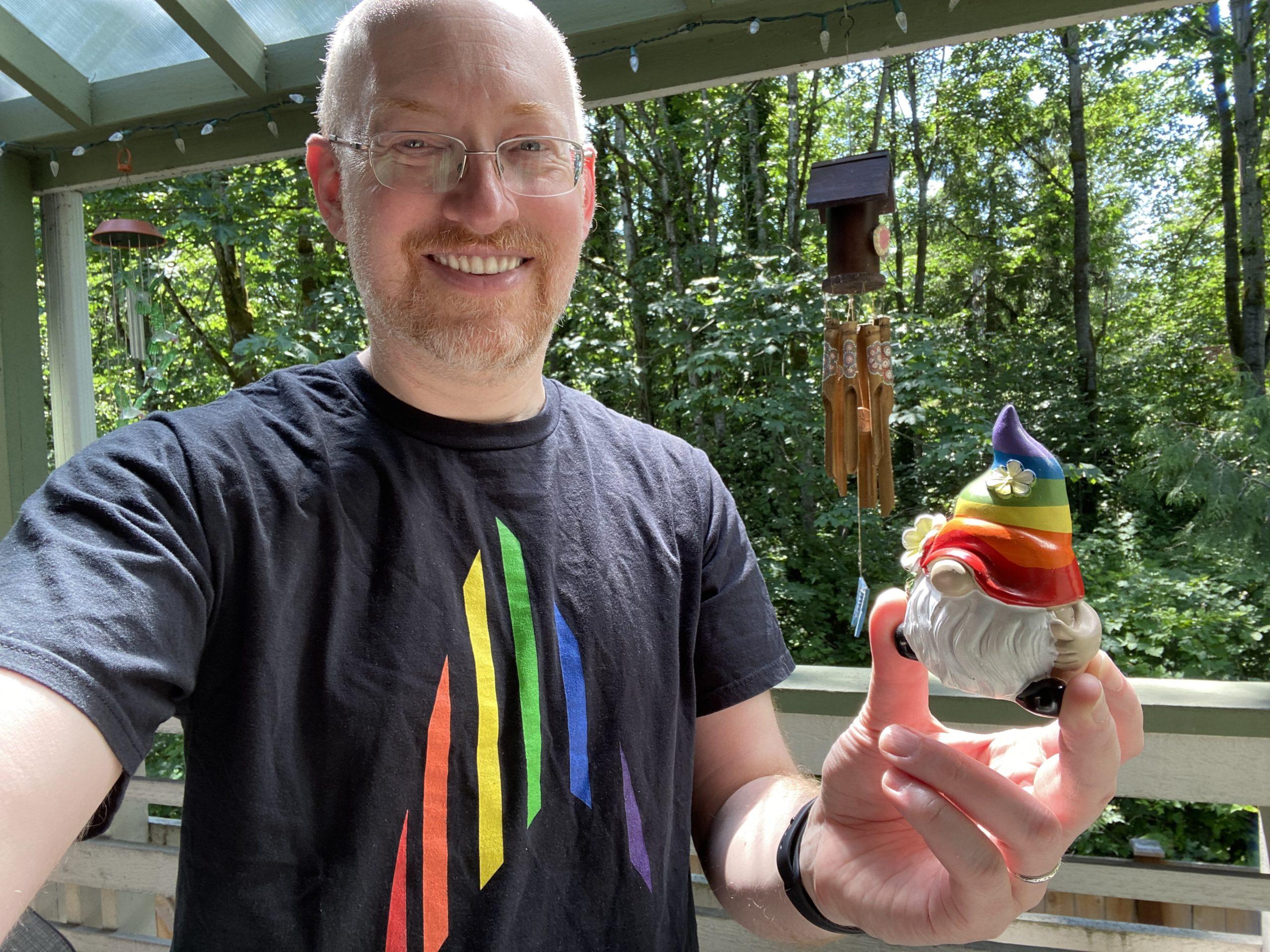 Me standing on my back deck with green trees in the background, wearing a black t-shirt with the Star Trek delta made of rainbow stripes, holding a small garden gnome sculpture; the gnome is wearing a rainbow striped hat.