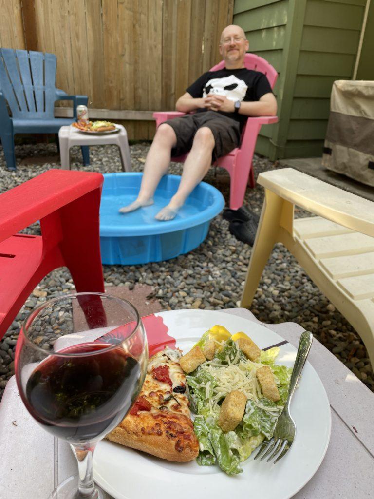 In the foreground is a half-glass of red wine next to a plate with pizza and salad. In the background, slightly out of focus, I’m sitting in a pink plastic Adirondack chair in our back yard, next to my own dinner plate and drink on a small plastic table, with my feet submerged in a blue plastic kiddie pool full of water.