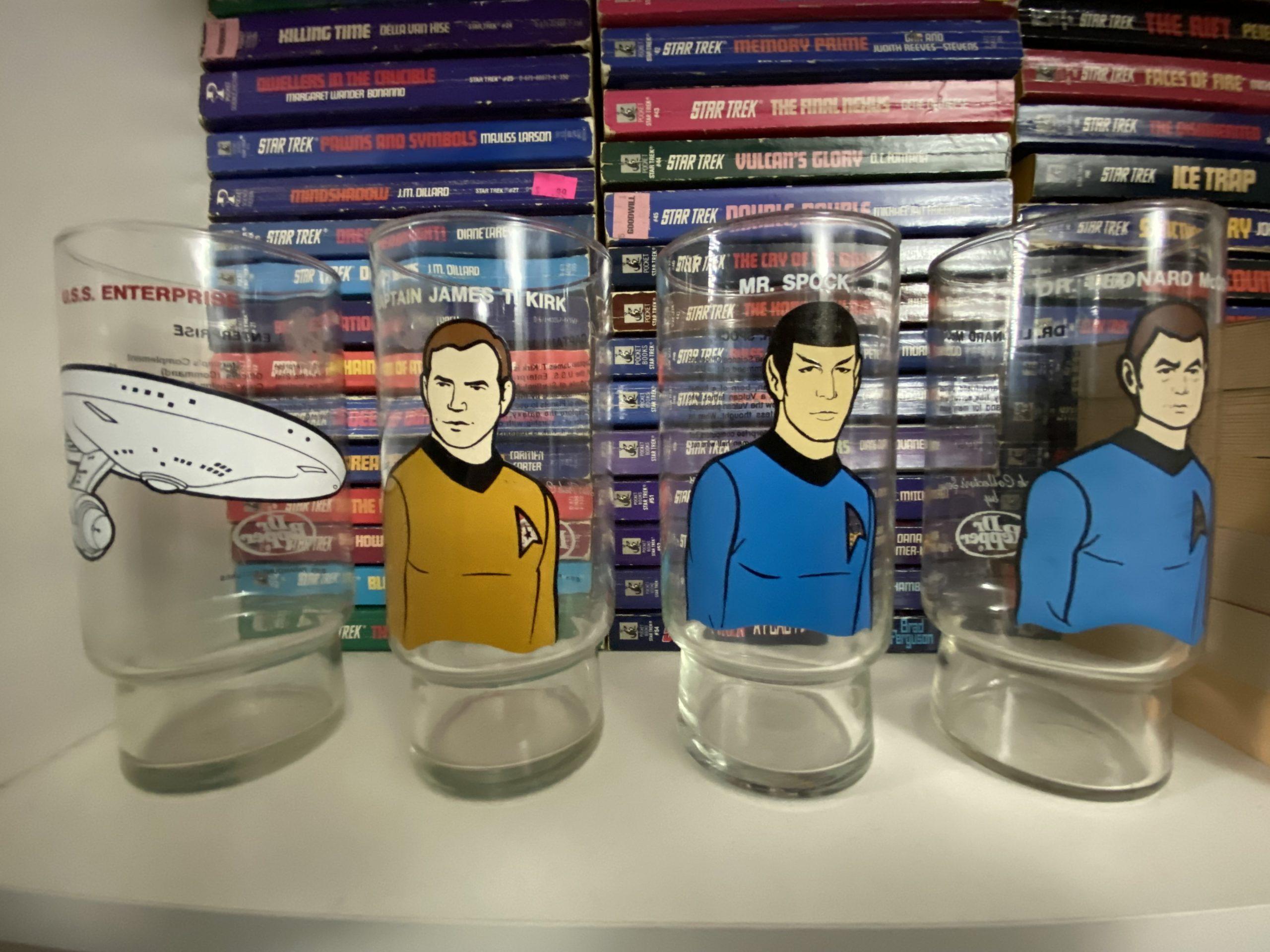 Four drinking glases sitting on a bookshelf in front of a stack of TOS Star Trek novels. TAS artwork on the glasses shows the Enterprise, Kirk, Spock, and McCoy.