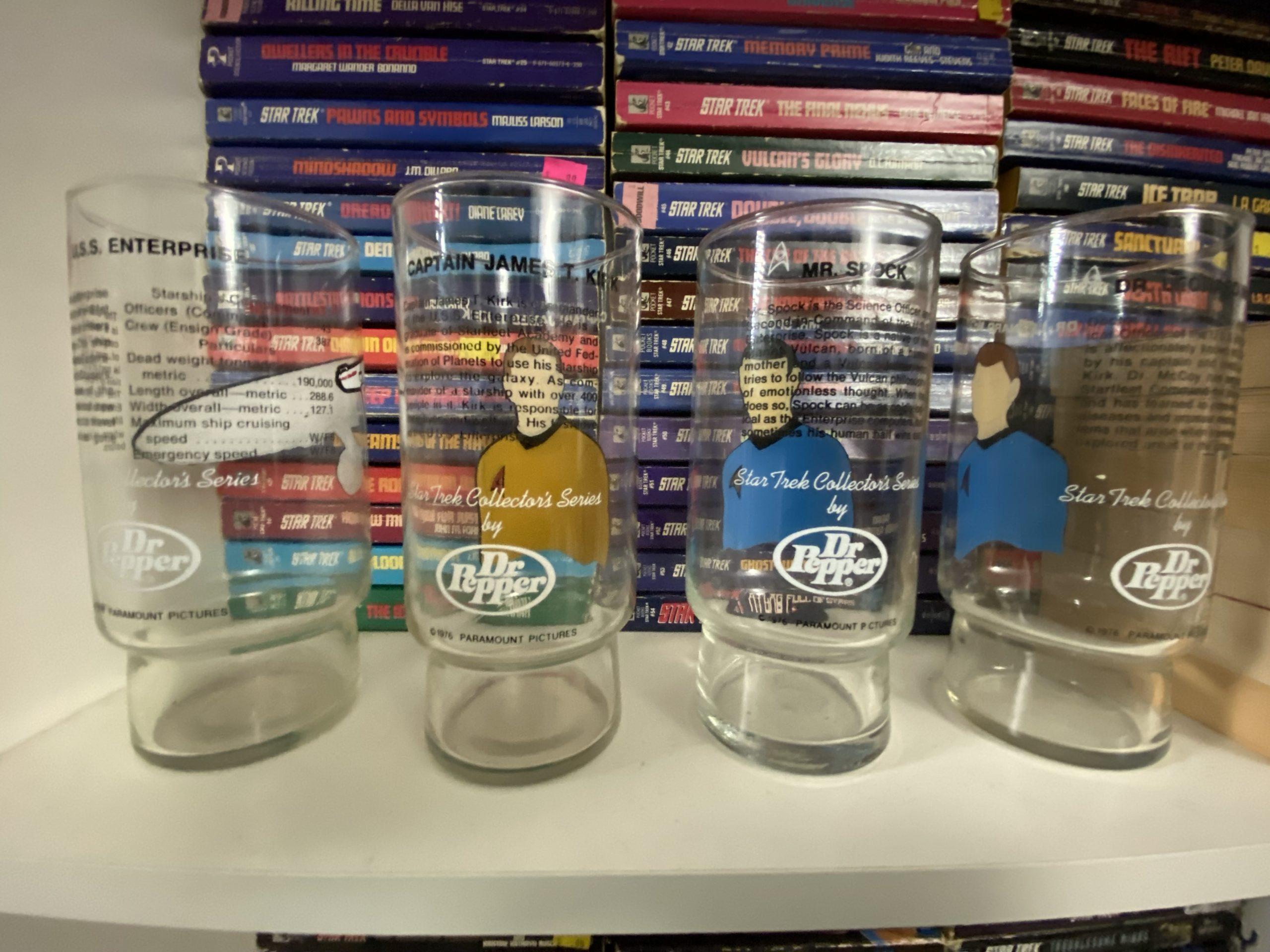 Four drinking glases sitting on a bookshelf in front of a stack of TOS Star Trek novels. They are turned to show the text on the back of each glass, though it's difficult to read in this photo.