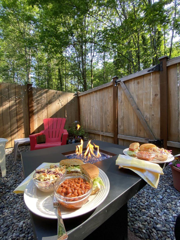 A wide angle shot of our backyard, with tall trees beyond the fence, and a fire pit table with our dinner plates in the foreground.