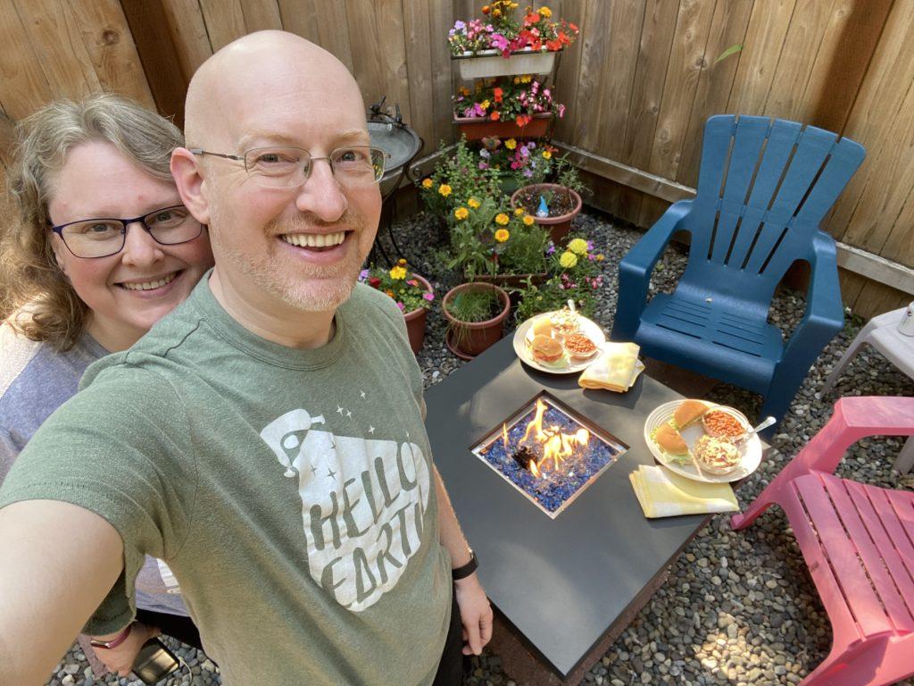 My wife and I in our backyard, in front of a small fire pit table with plates of burgers, coleslaw, and beans.