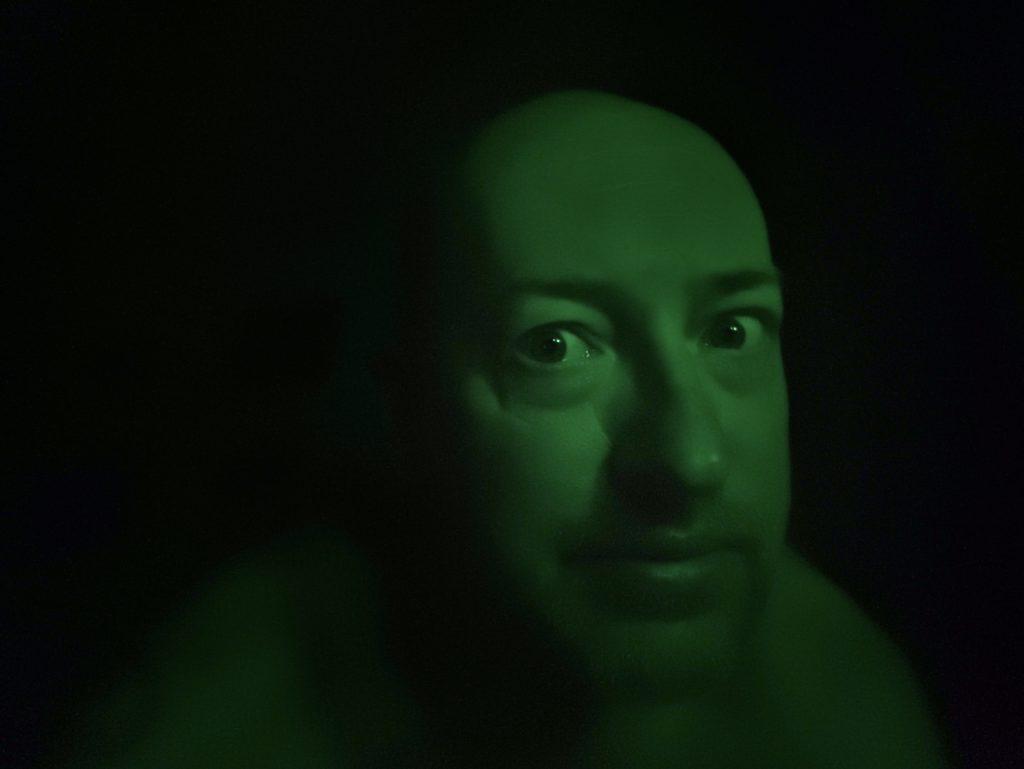 A very dark selfie, lit only in green; only my face is visible against a black background.