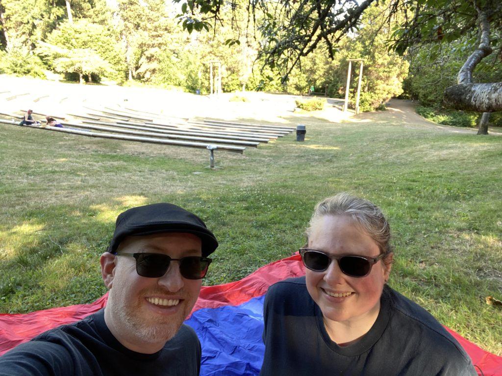 My wife and I sitting outdoors in a park on a blue and red picnic blanket; behind us is a mostly empty amphitheater and stage being prepped for a play.