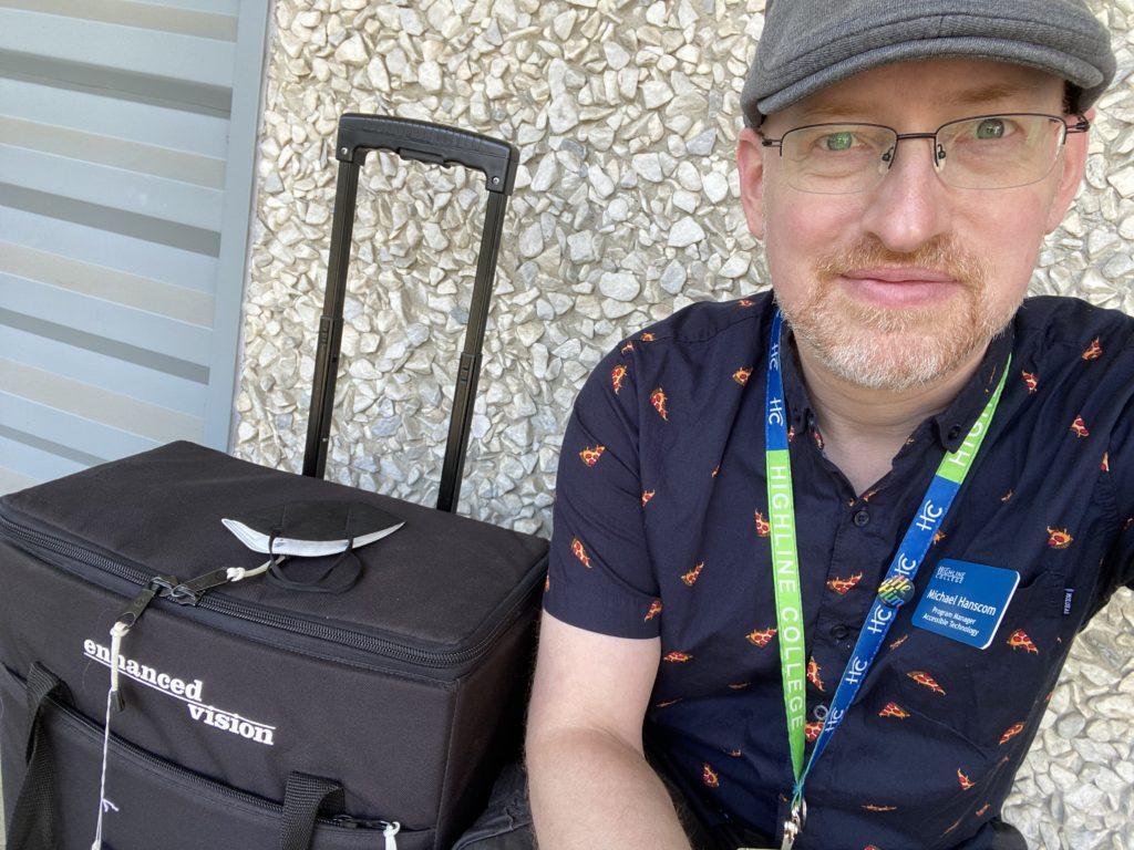 Me crouching in front of a 60s-era pebbled building wall, next to a black padded rolling case about the size of carry-on luggage labeled ‘Enhanced Vision’. I’m wearing a grey cap and blue shirt with a print of pizza slices.