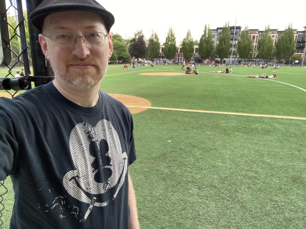 Me, wearing a black t-shirt with a print of Mickey Mouse smoking a cigarette, standing in front of a park playfield with a lot of people sitting in small groups enjoying the summer evening weather.