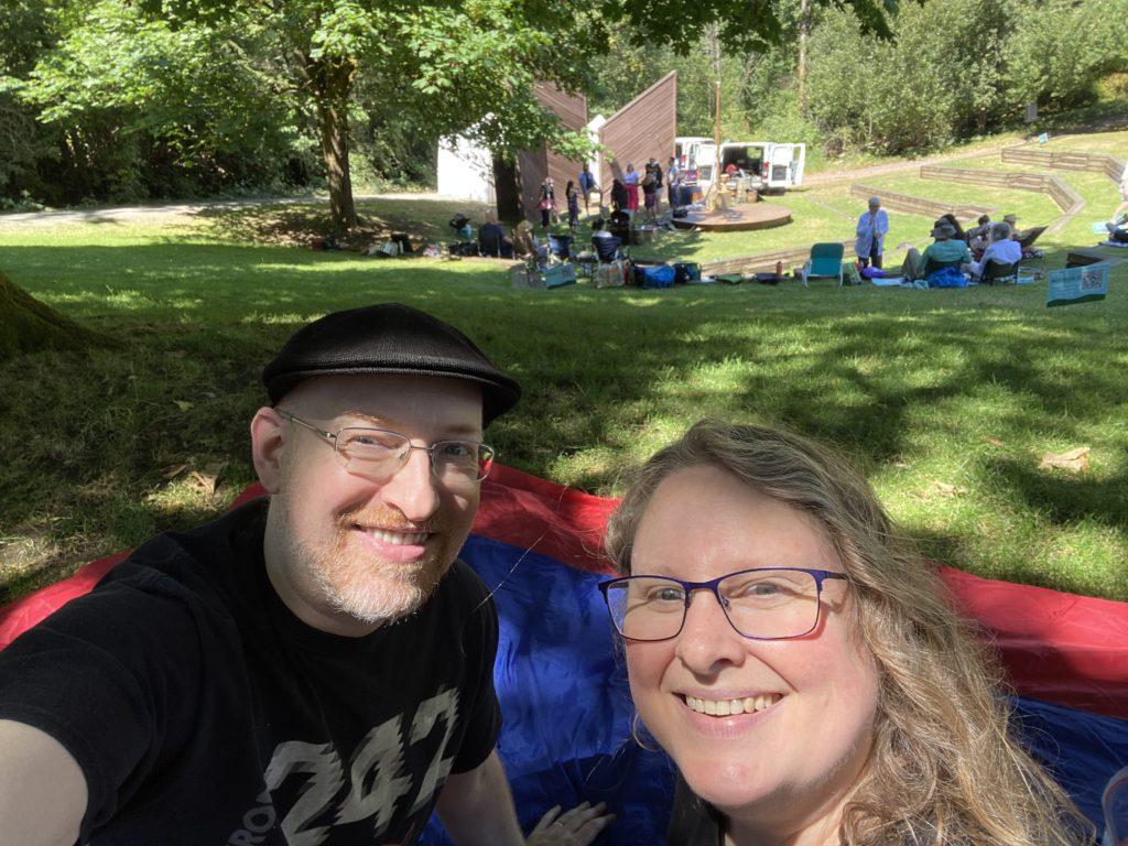 My wife and I in a sunny park, sitting in the shade under a tree, with a park amphitheater in the background being prepped for a performance.