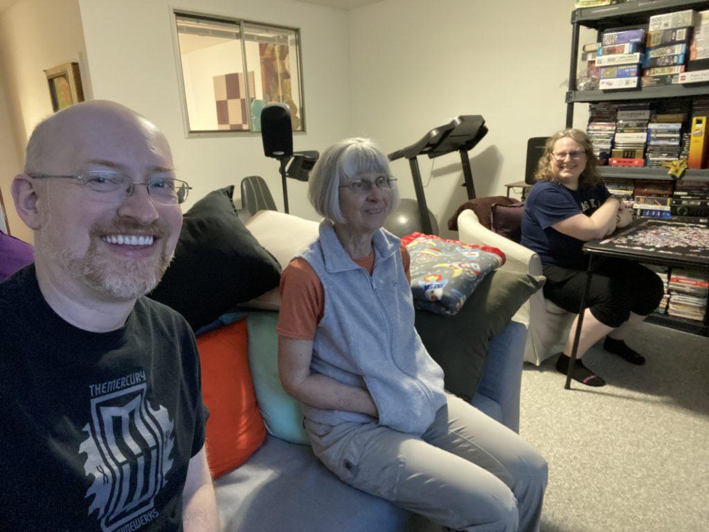 Me, my mother-in-law, and my wife sitting in our basement. I and my MIL are sitting on a couch, my wife is at a card table in front of a just-started puzzle. A shelf full of movies and more puzzles is behind her.