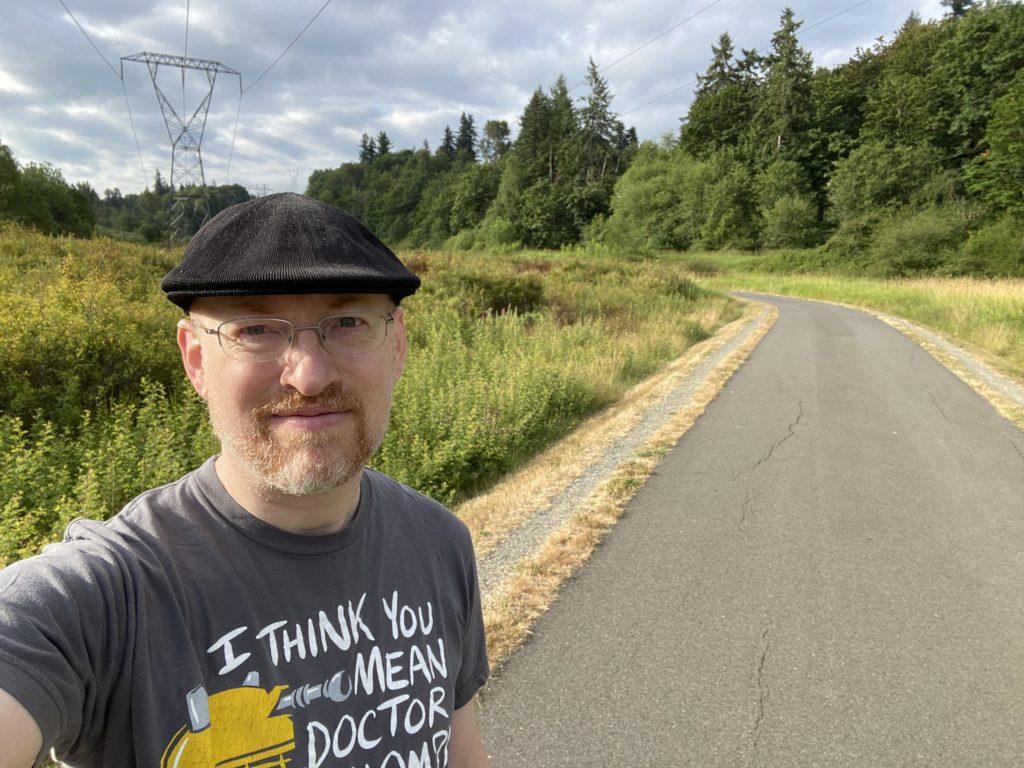 Me standing outdoors on a paved path through a grassy clearing with trees to the sides and in the background. A power line tower is behind me. The sky is cloudy, but sun is shining where I’m standing.