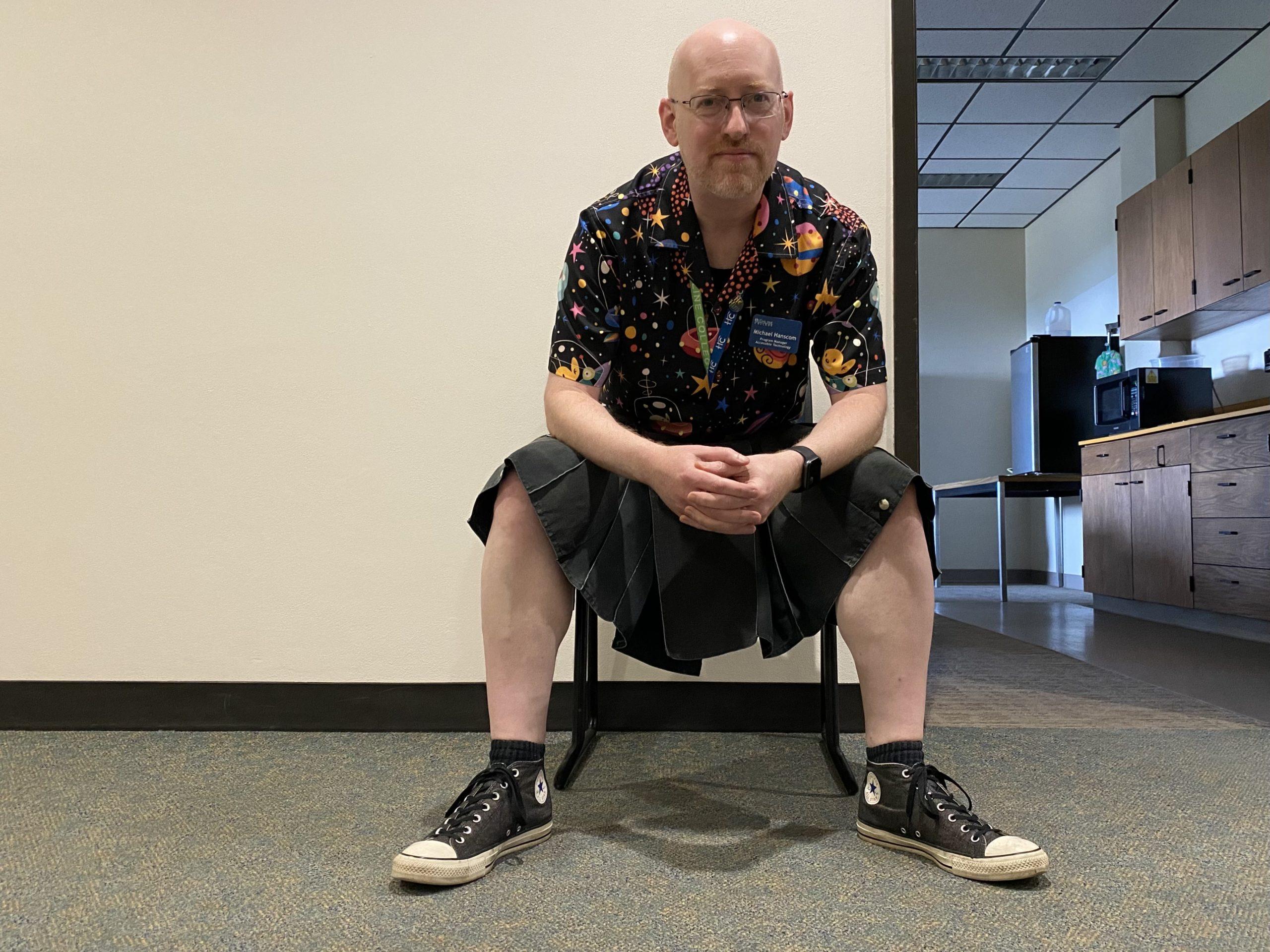 Me sitting on a chair in a hallway next to an open door to a breakroom. I'm leaning forward with my arms on my knees, wearing grey converse, a black kilt, and a black button-up short-sleeve shirt with a pattern of cartoon aliens.