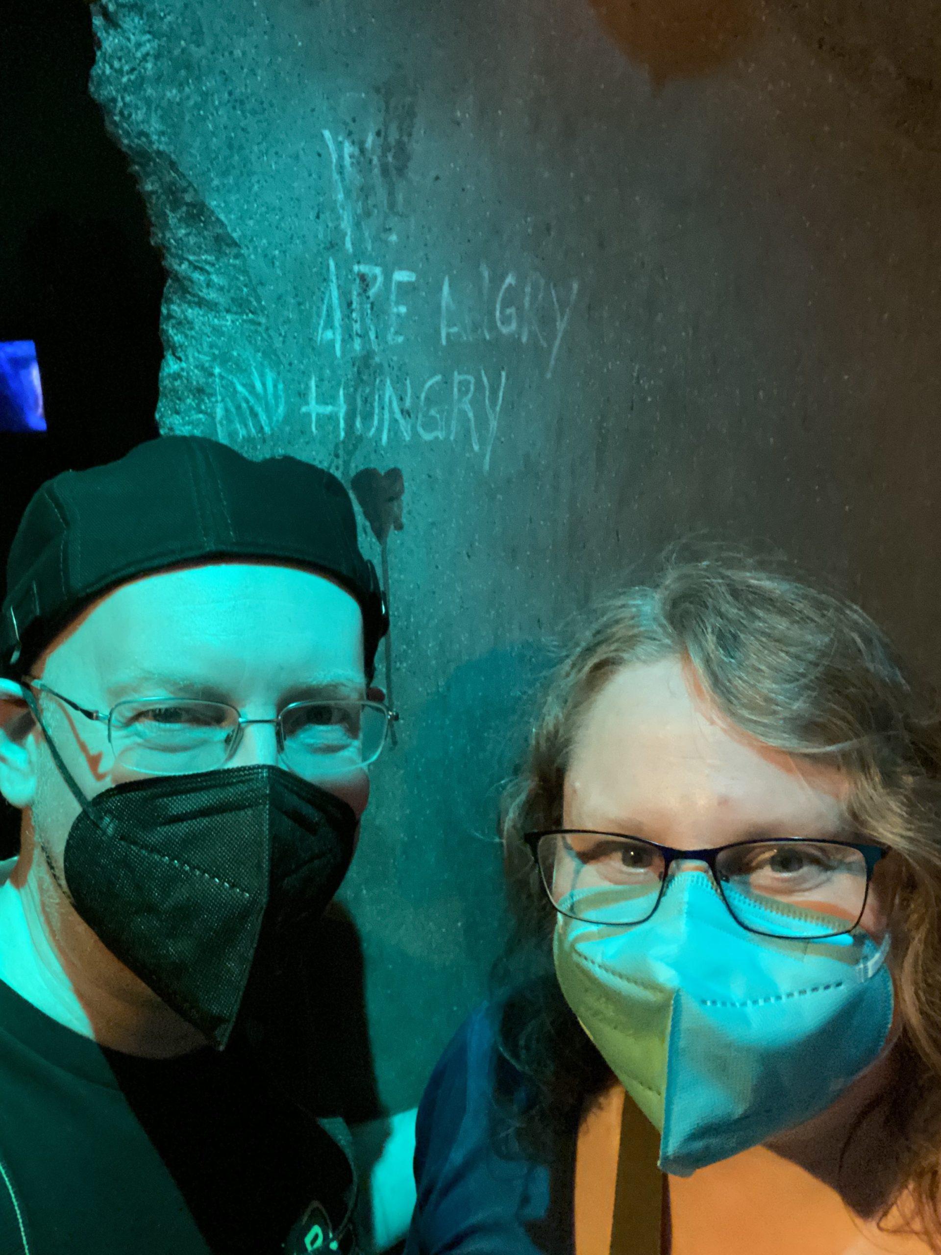 My wife and I, both wearing face masks, standing in front of a blue-lit concrete wall with the words "we are angry and hungry" scratched into it.