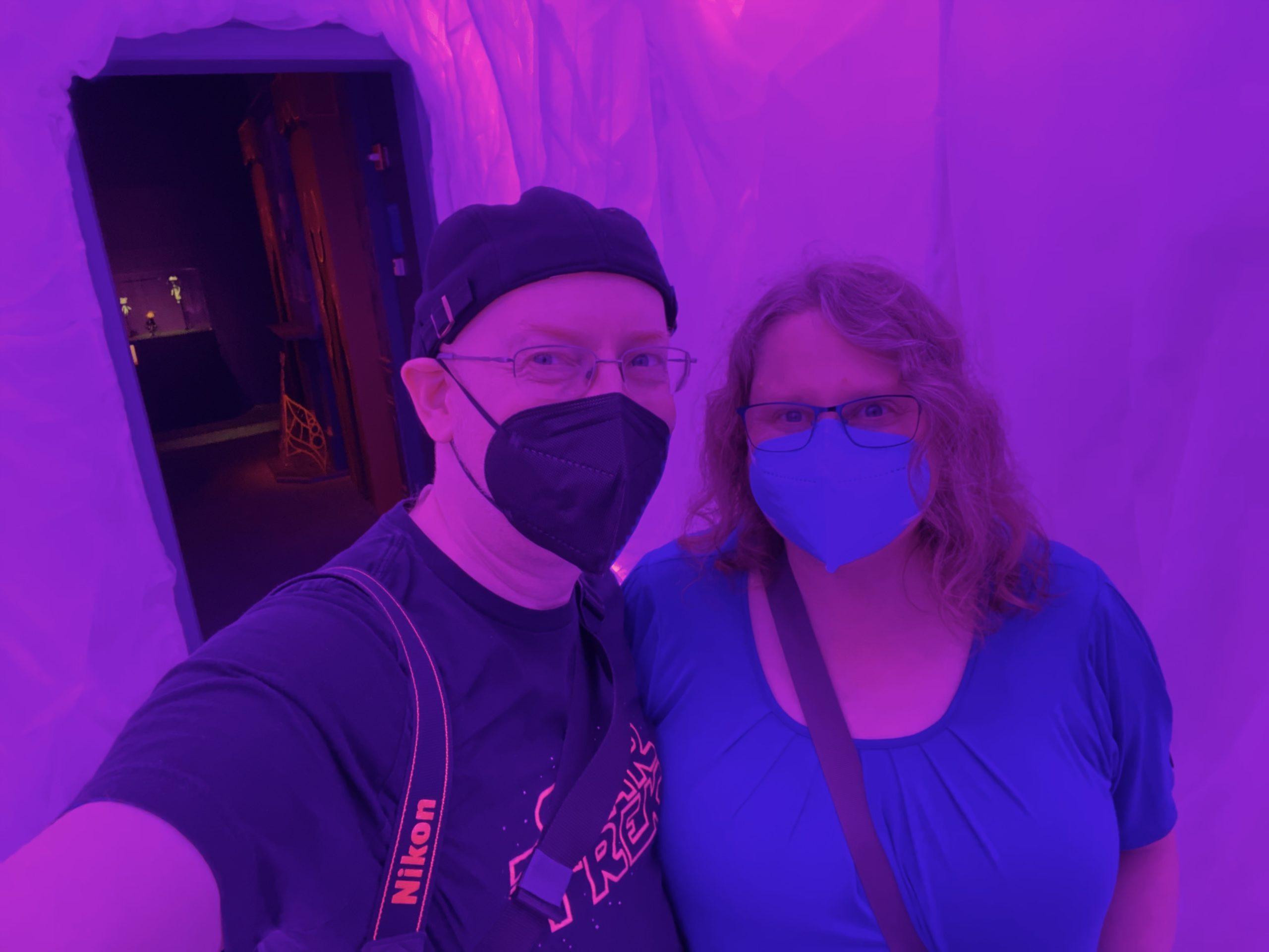 My wife and I, both wearing face masks, in a very pink hallway, lit in pink, so the picture has a very pink cast to it.