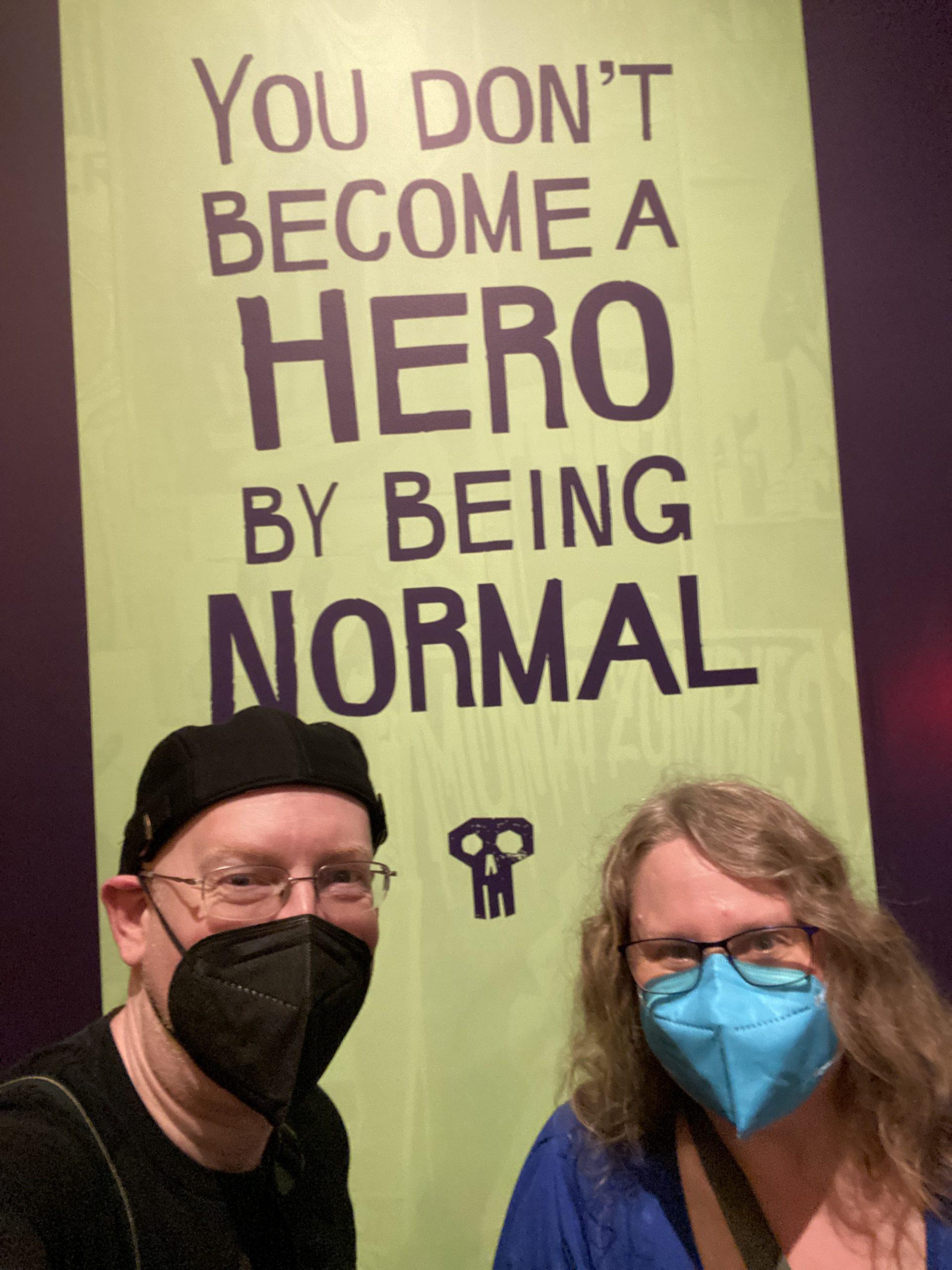 My wife and I in front of a sign saying "you don't become a hero by being normal".