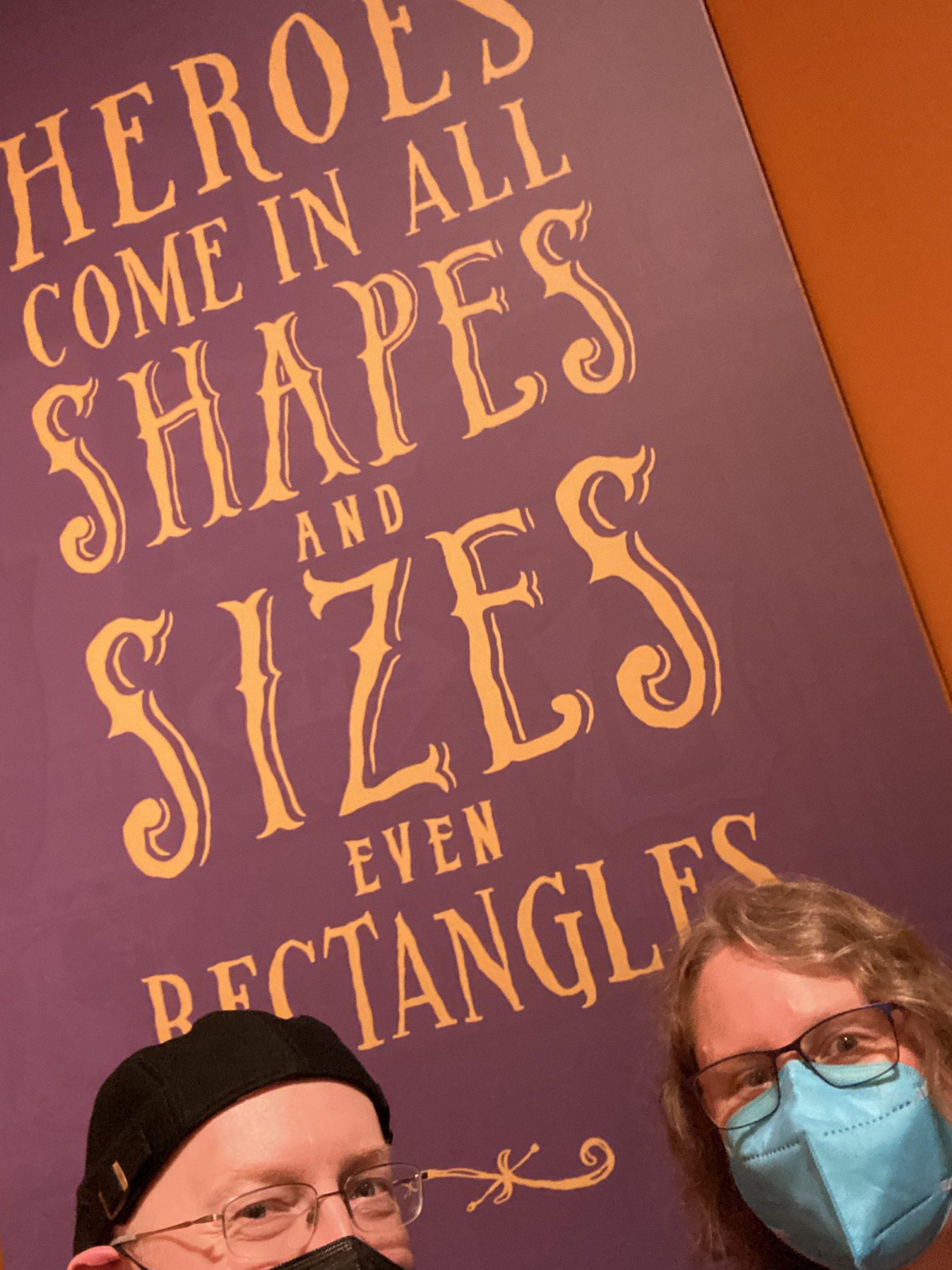 My wife and I in front of a sign saying "heroes come in all shapes and sizes even rectangles".